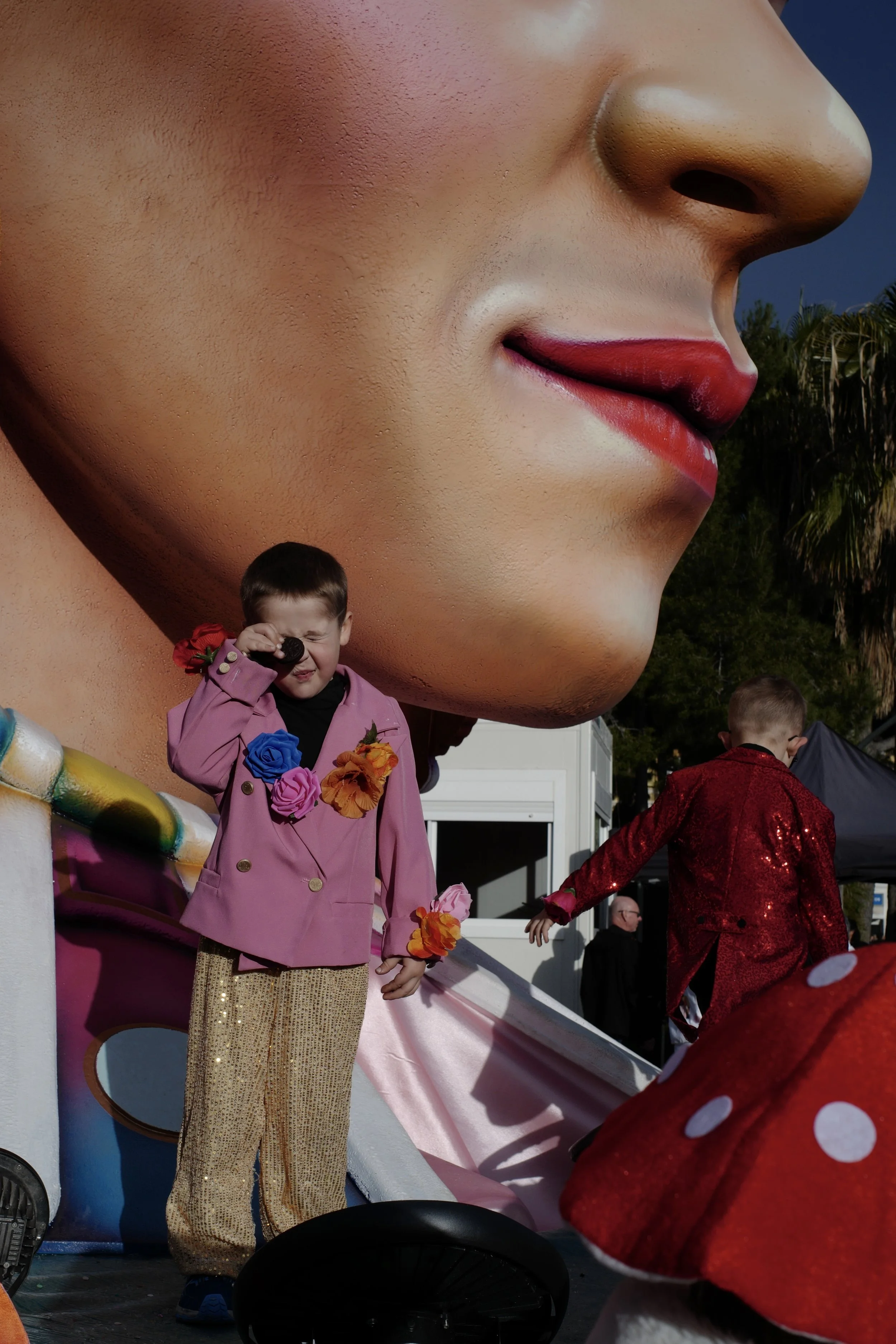 A boy in a pink jacket and gold pants holds a brown cookie to his right eye, covering it. He stands in front of a giant profile of a face with red lips.