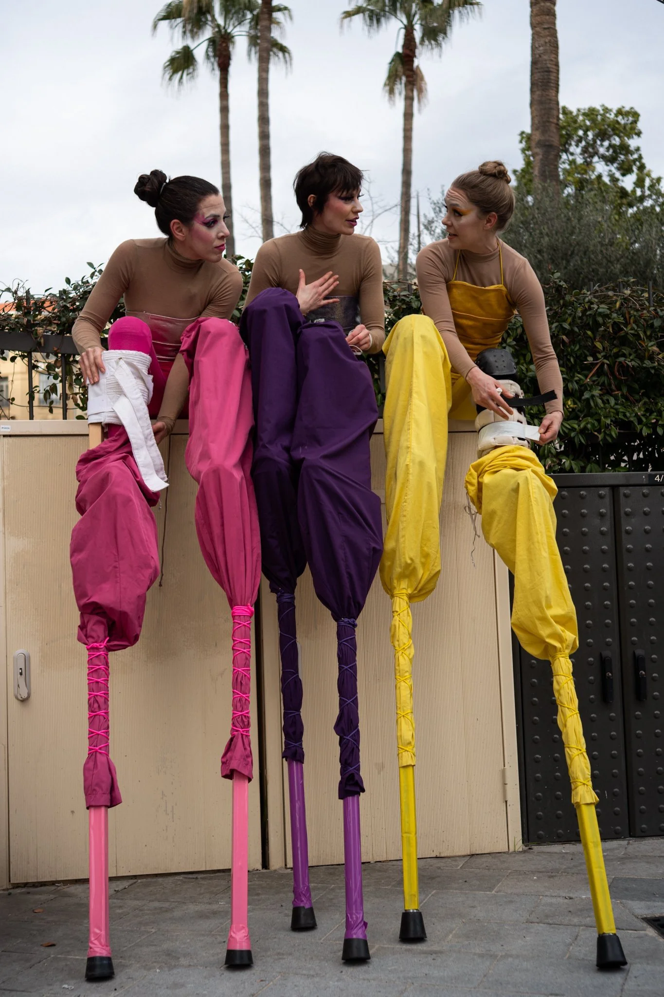 Three women on stilts sit atop a light-colored ledge, talking. They wear beige tops, with pink, purple, and yellow stilt covers. Palm trees are visible in the background under a cloudy sky.