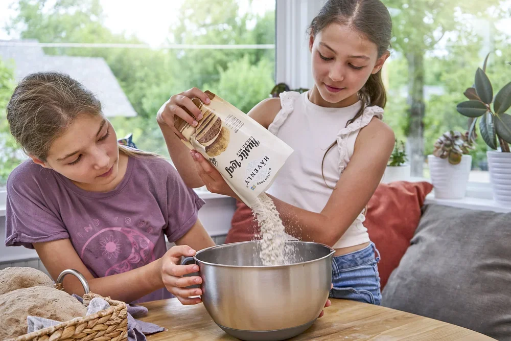 Two young girls baking together in a bright, cozy kitchen, one pouring flour into a mixing bowl.