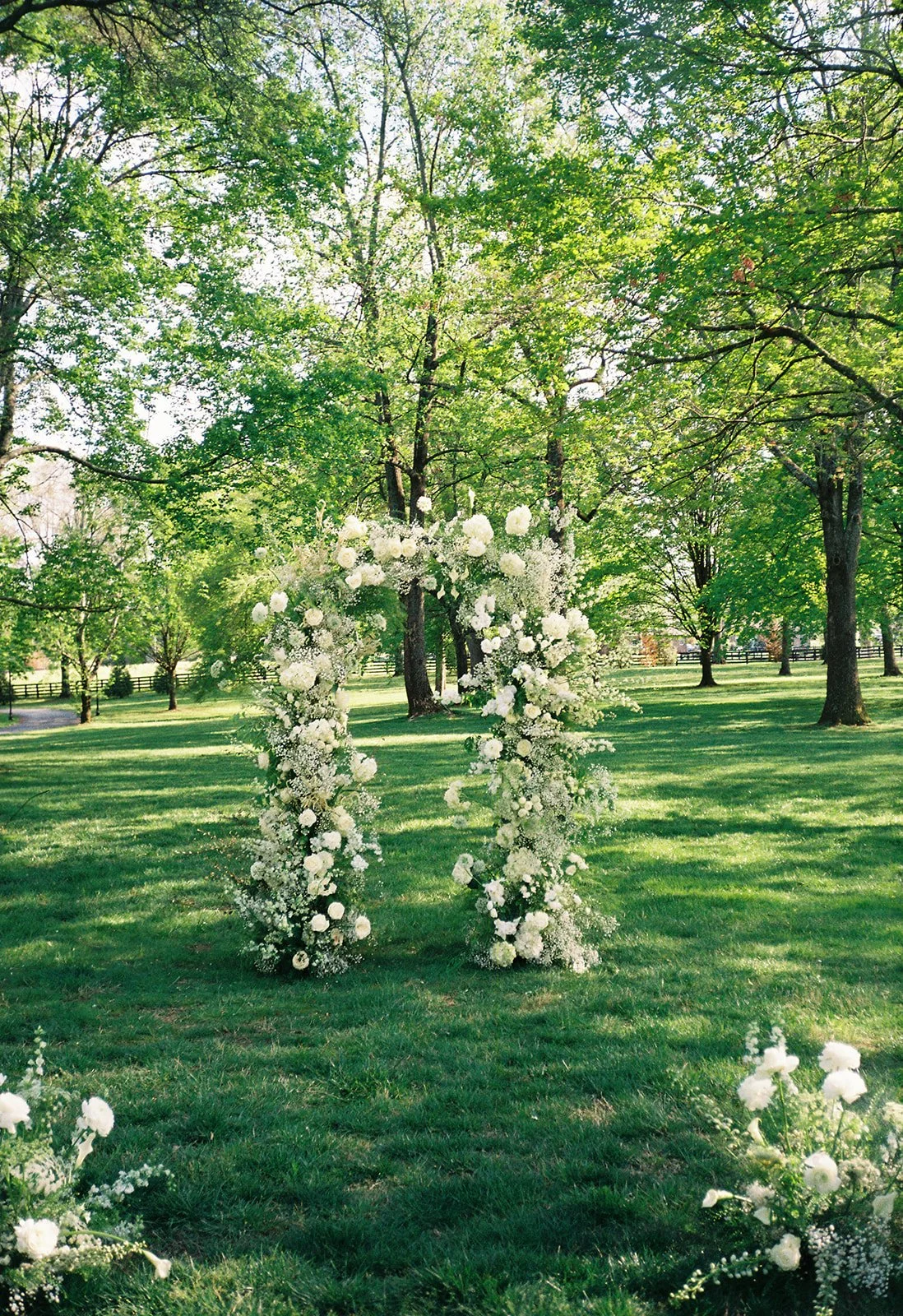 A floral arch decorated with white flowers standing in a grassy park area with green trees and a bright sky in the background.