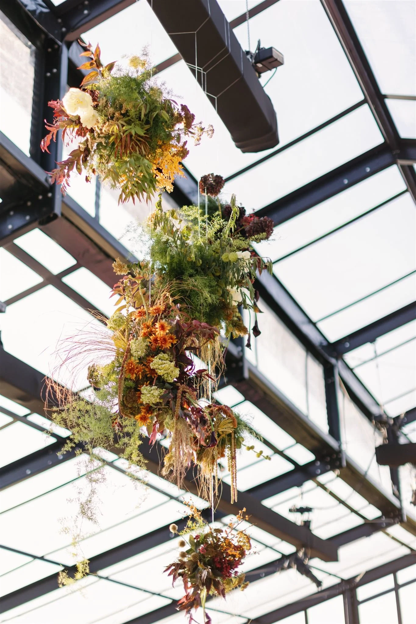 Hanging floral arrangement featuring various colorful flowers and greenery inside a glass conservatory or greenhouse.