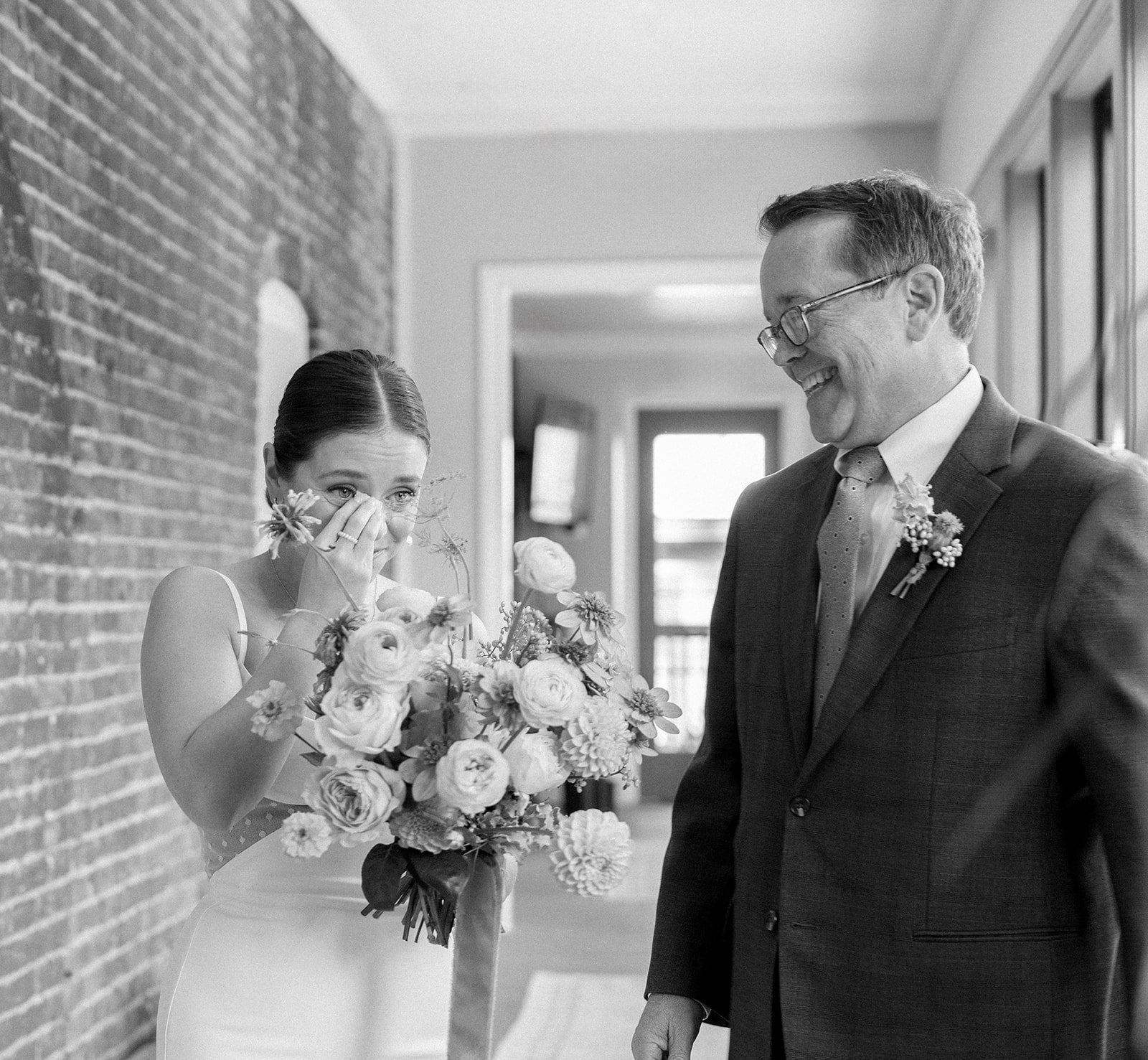 A bride with a bouquet of flowers wiping away tears as a smiling man in a suit, possibly the groom or father of the bride, looks at her.