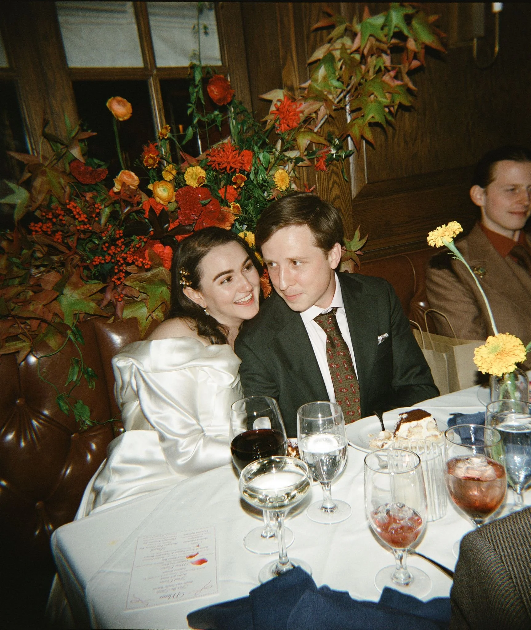 A woman in a satin dress and a man in a suit sitting at a dinner table with drinks and dessert, surrounded by floral arrangements, in a wood-paneled room.
