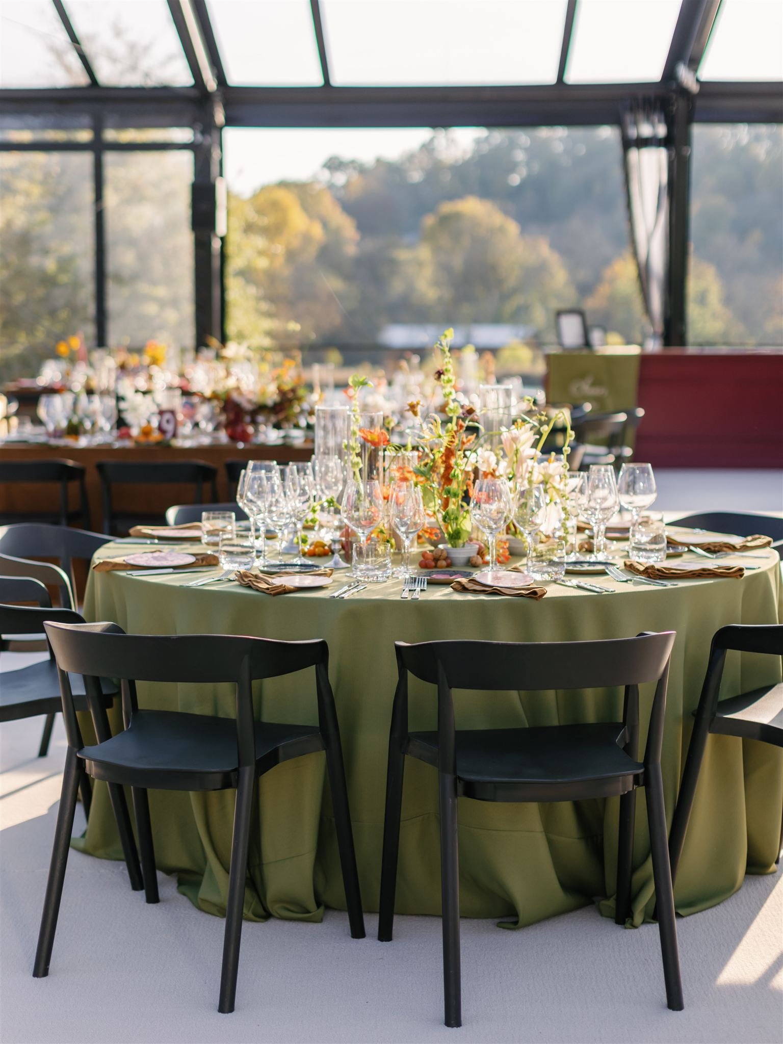 Round banquet table with a green tablecloth, set with wine glasses, water glasses, cutlery, salt and pepper shakers, napkins, and a floral centerpiece, in a sunlit space with large windows showing trees outside.