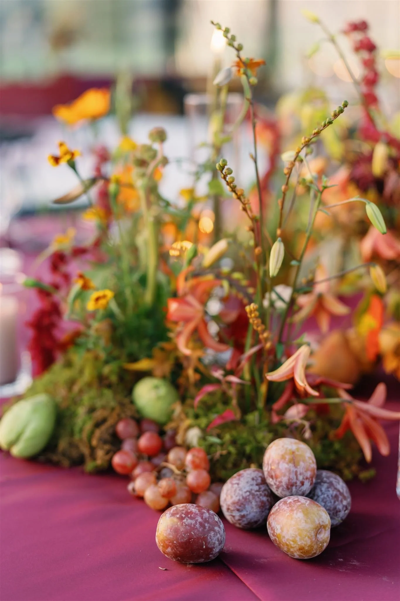 A floral arrangement with various flowers, grapes, and decorative elements on a table, set in a softly lit indoor environment.