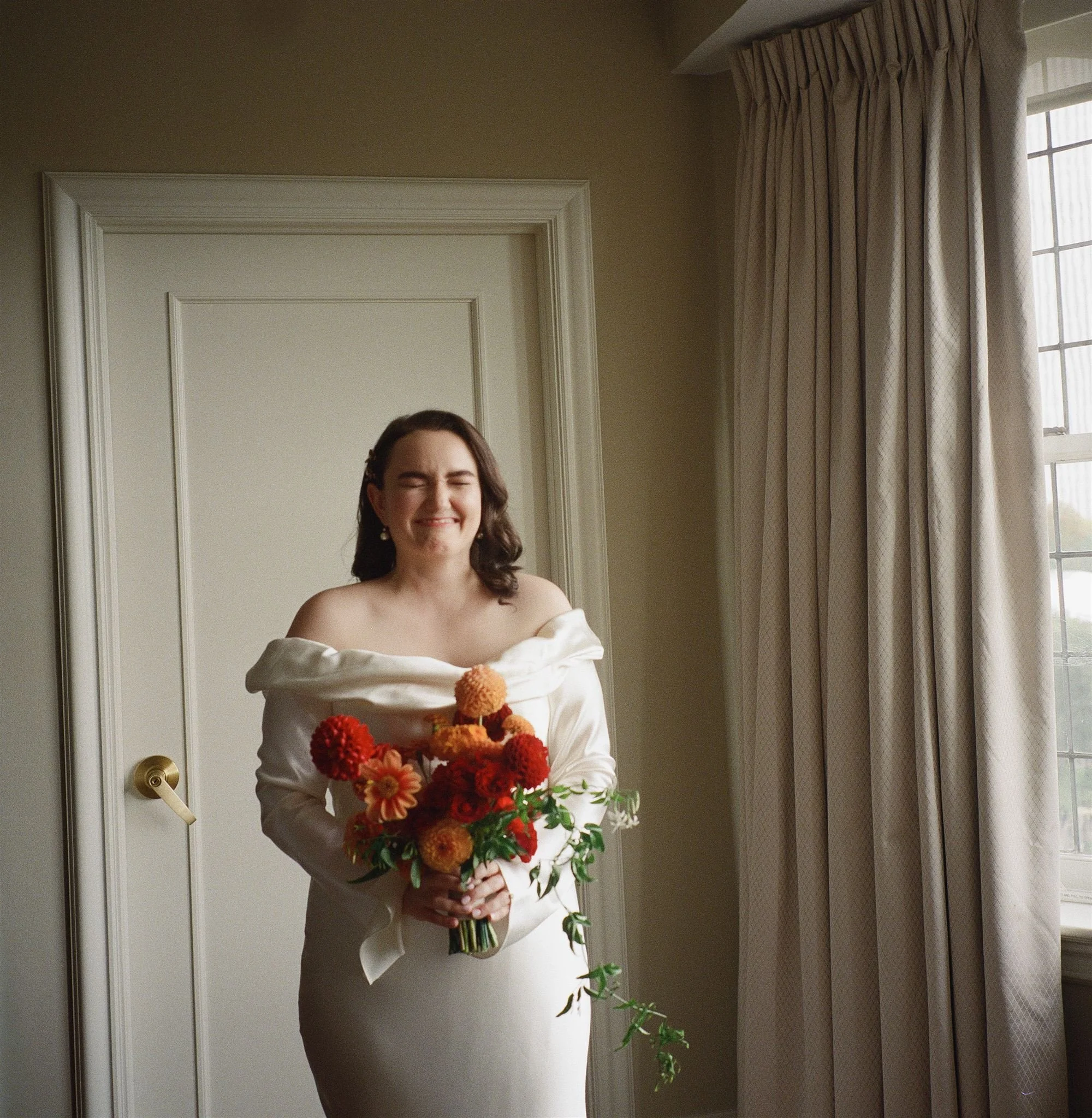 A woman in a white off-shoulder dress holding a bouquet of red and orange flowers, standing in a room with a cream-colored door and beige curtains by a window.