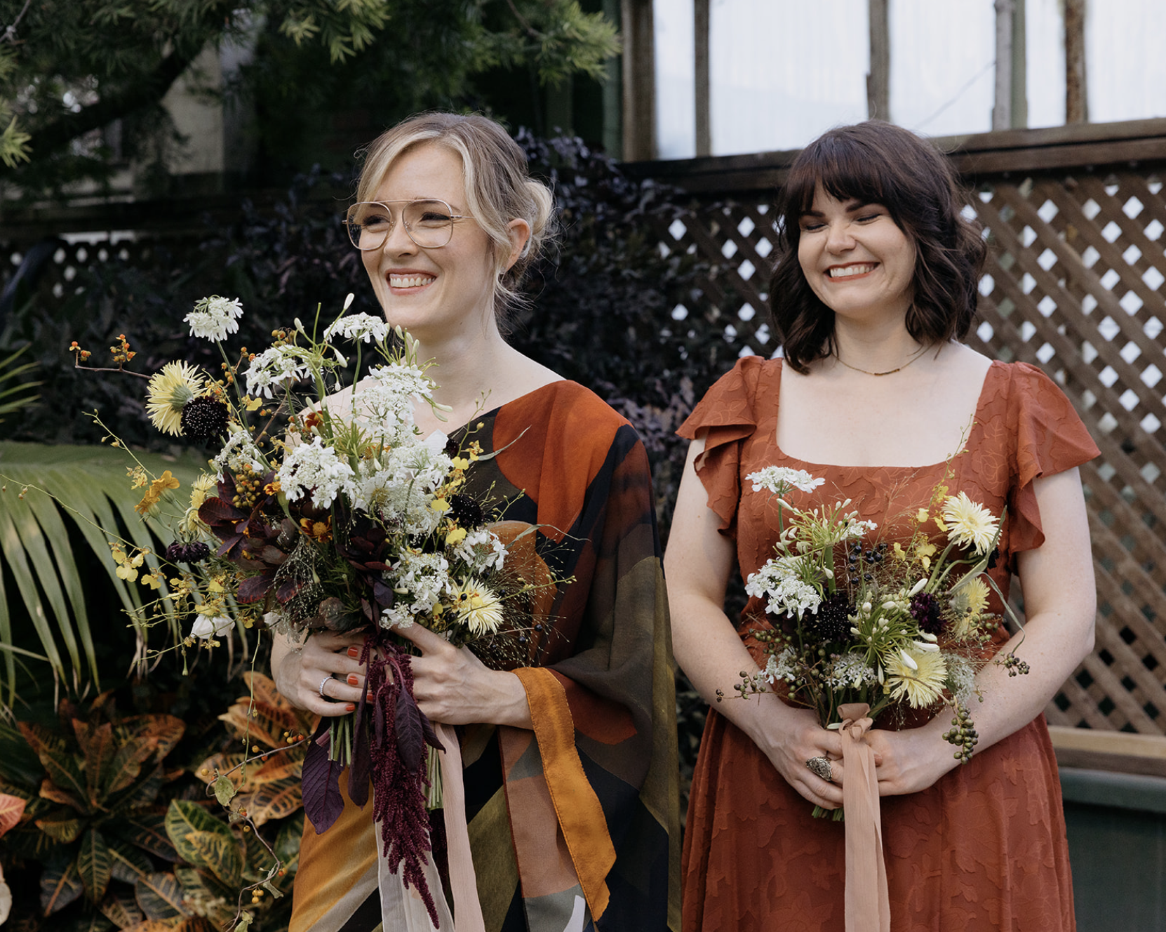 Two women smiling and holding bouquets of flowers outdoors, with a wooden fence and greenery in the background.