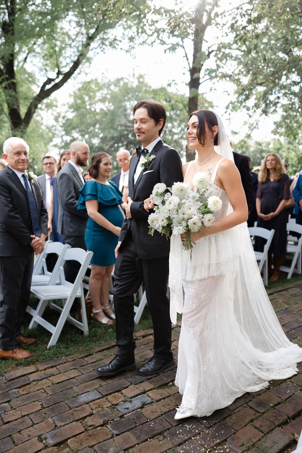 Bride and groom walking down the aisle during their outdoor wedding ceremony, with guests standing on either side under trees.