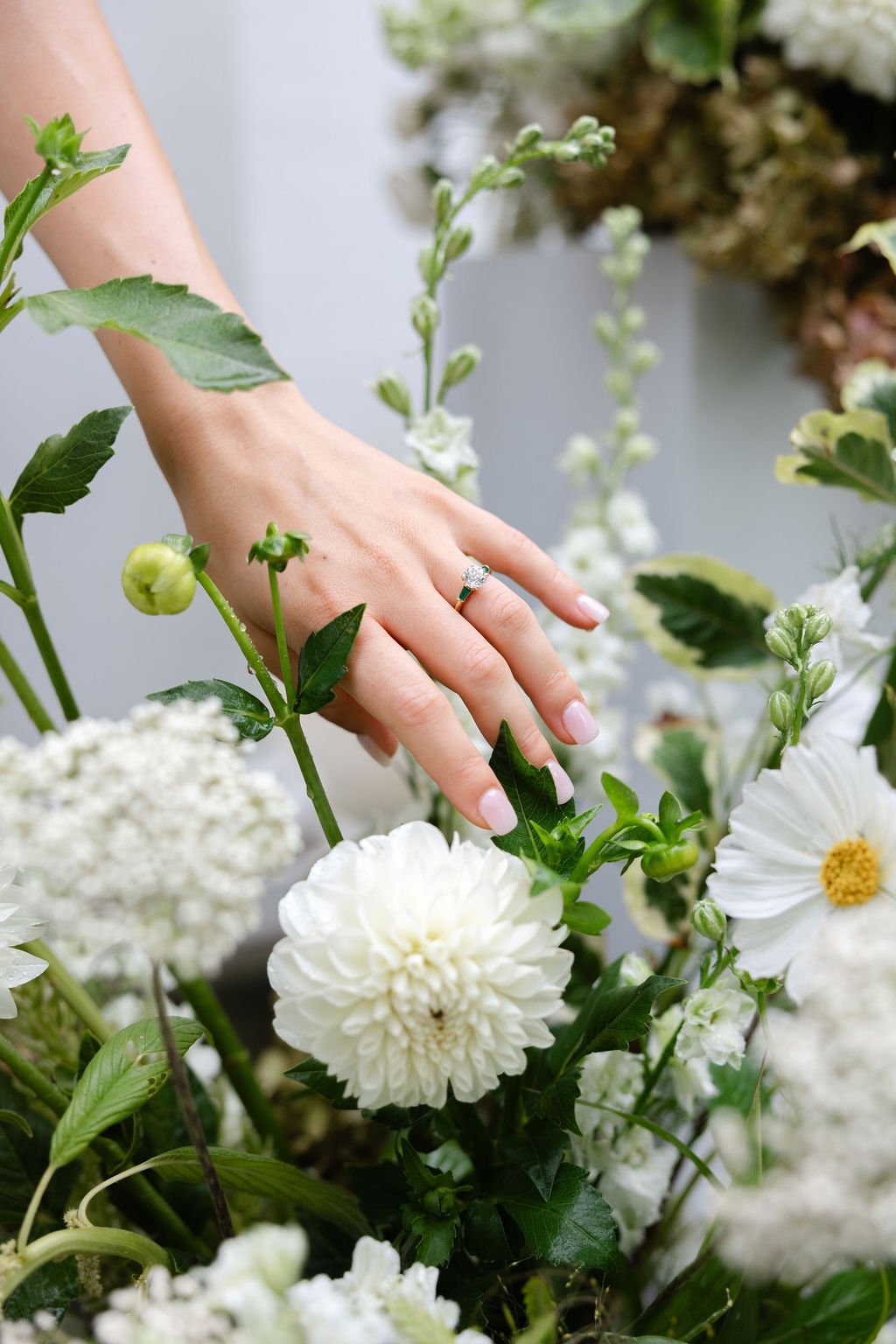A woman's hand with a diamond ring touching white flowers and green foliage.