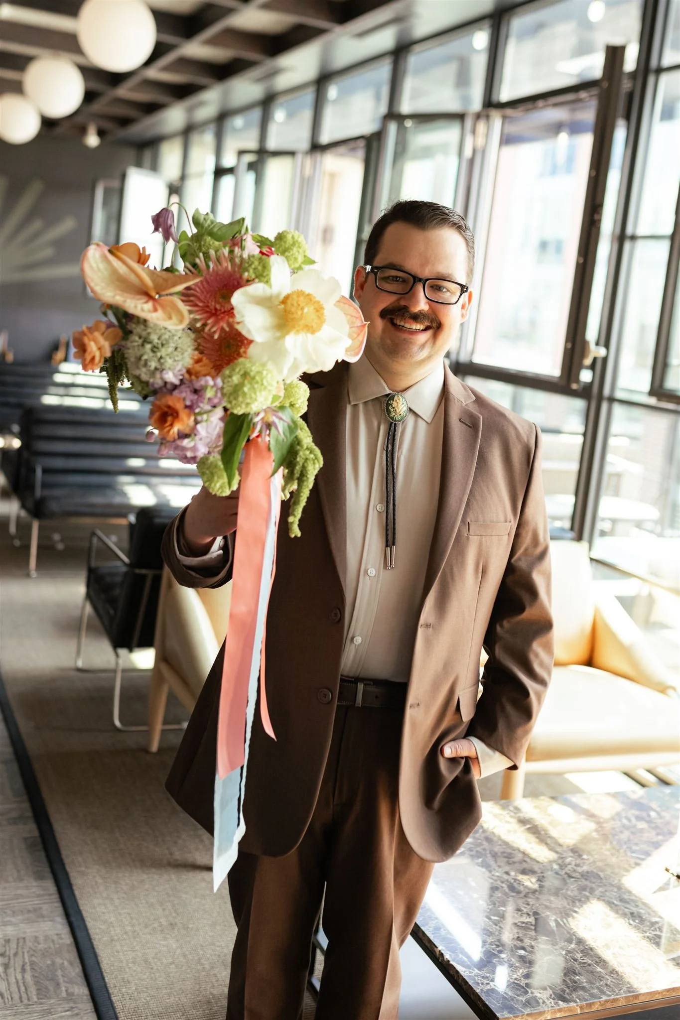 A man in a tan suit holding a colorful bouquet of flowers inside a modern indoor setting with large windows.