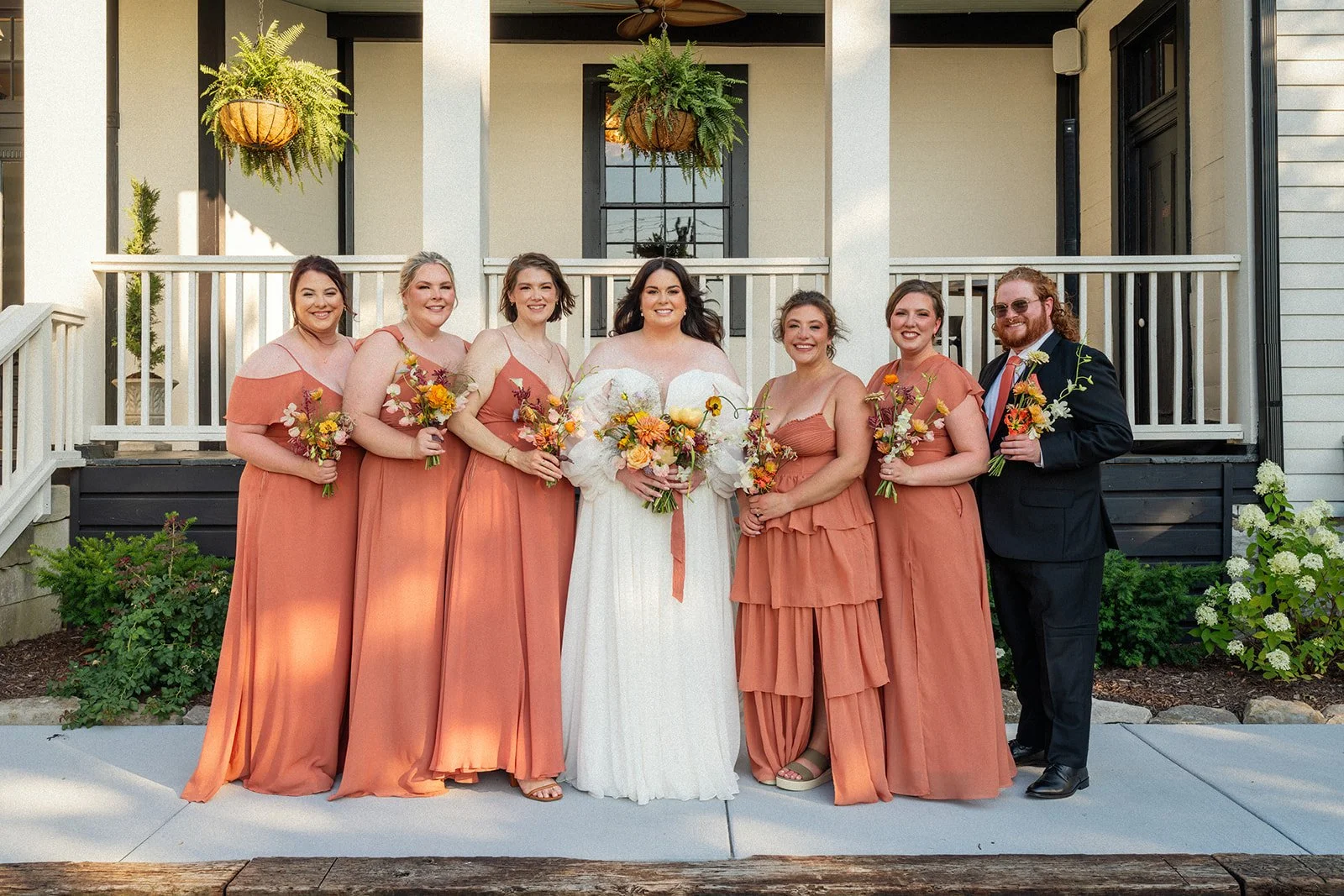 Group of seven women in peach dresses and one man in a suit, standing outside a house, with the woman in white holding a bouquet, during a wedding.