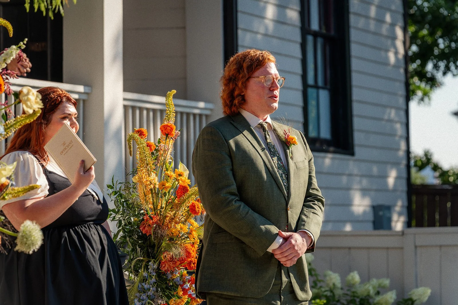 A man in a gray suit with a floral tie and glasses stands outdoors with his hands clasped, during a wedding ceremony. A woman with red hair and a white blouse with puffy sleeves holds a wedding program and stands near a large basket of orange and yel