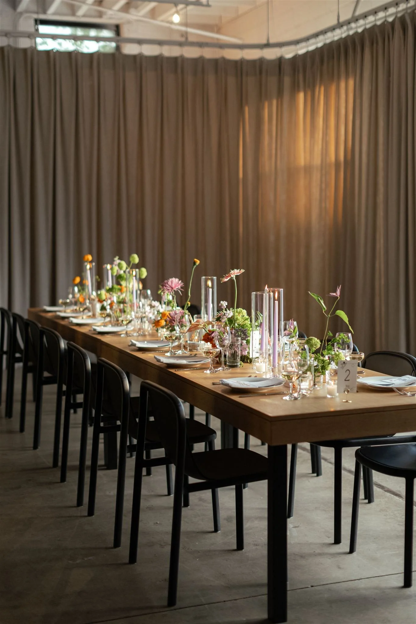 Long wooden dining table decorated with flowers and candles, set for a formal event in a room with beige curtains.