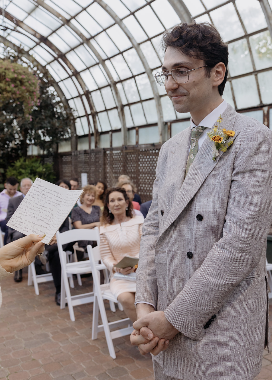 A man in a light gray suit with a boutonniere of yellow flowers stands with his hands clasped, during a wedding ceremony in a greenhouse-like setting. Guests seated behind him are watching, some holding programs.
