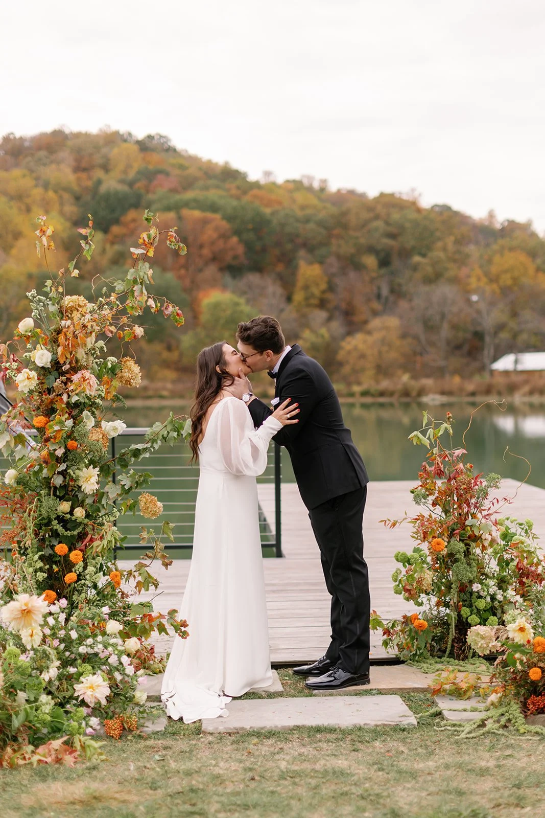 A bride and groom kissing during their outdoor wedding ceremony on a dock by a lake, surrounded by fall foliage and floral decorations.