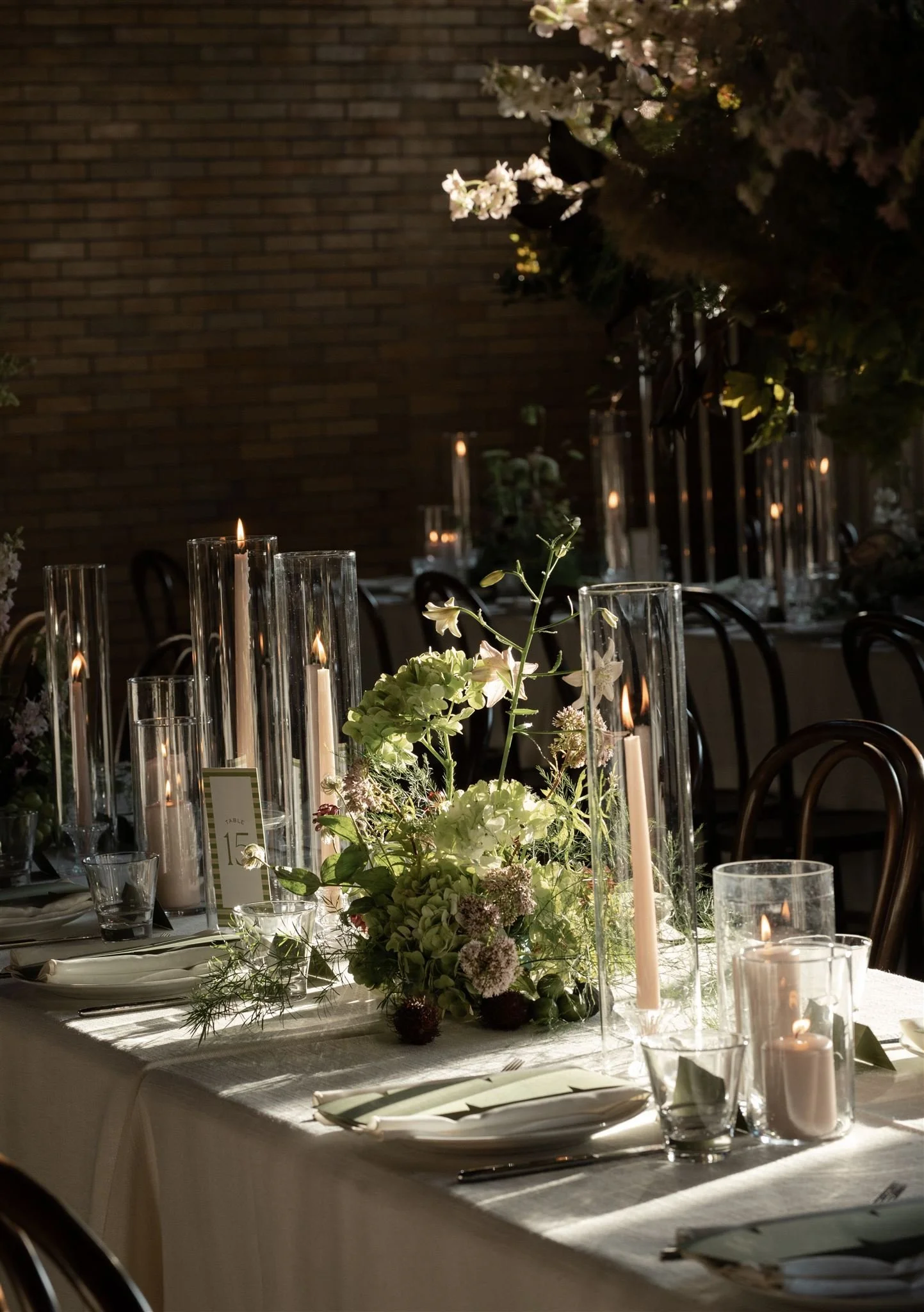 Elegant dinner table decorated with white and green floral centerpieces and tall glass candle holders with lit white candles, set with white plates, napkins, and cutlery, in a dimly lit setting with warm candlelight and a brick wall background.