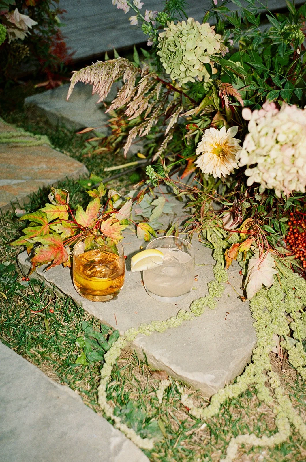 Two cocktails with lemon slices on a stone platform, surrounded by autumn leaves and flowers, outdoors.