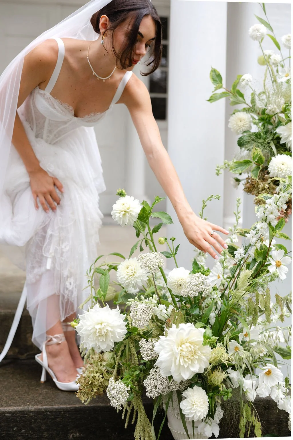 A bride in a white wedding dress adjusting her gown beside a large floral arrangement of white flowers and greenery.