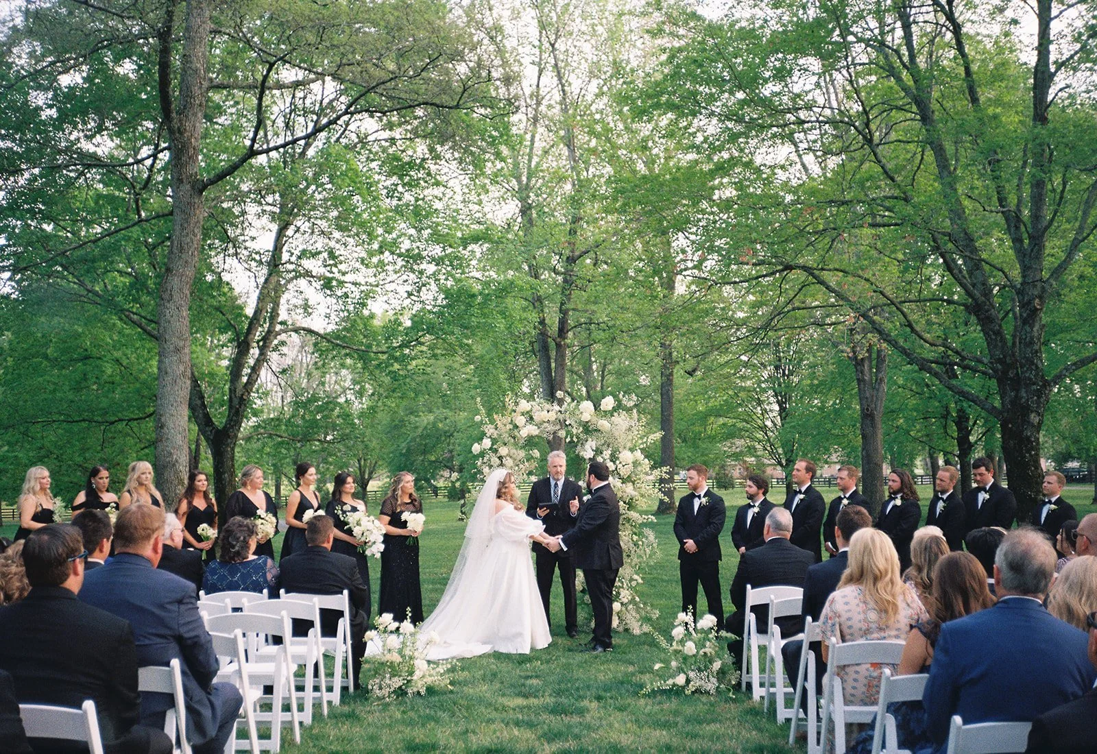 A wedding ceremony taking place outdoors in a park with green trees and grass. The bride and groom are standing in front of a floral arch, holding hands, with the officiant between them. Bridesmaids and groomsmen are lined up on either side, and gues