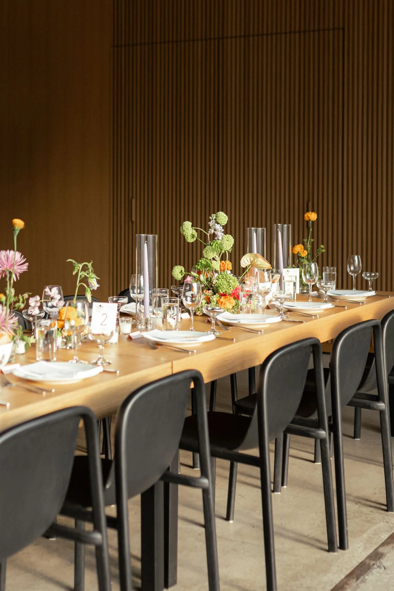 A long wooden dining table set for a formal event with floral centerpieces, glassware, plates, silverware, and black chairs in a modern interior with wood-paneled walls.