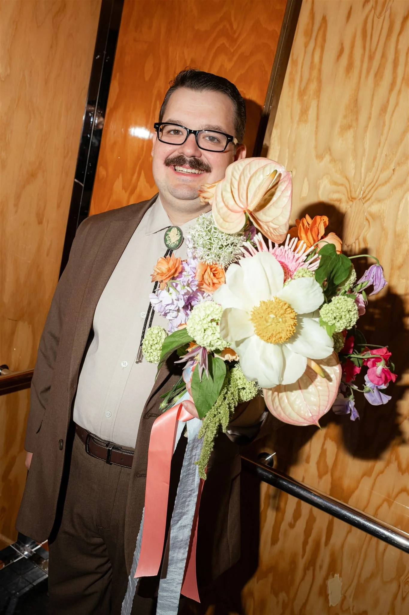 A man in a suit holding a colorful bouquet of flowers in an elevator with wood-paneled walls.