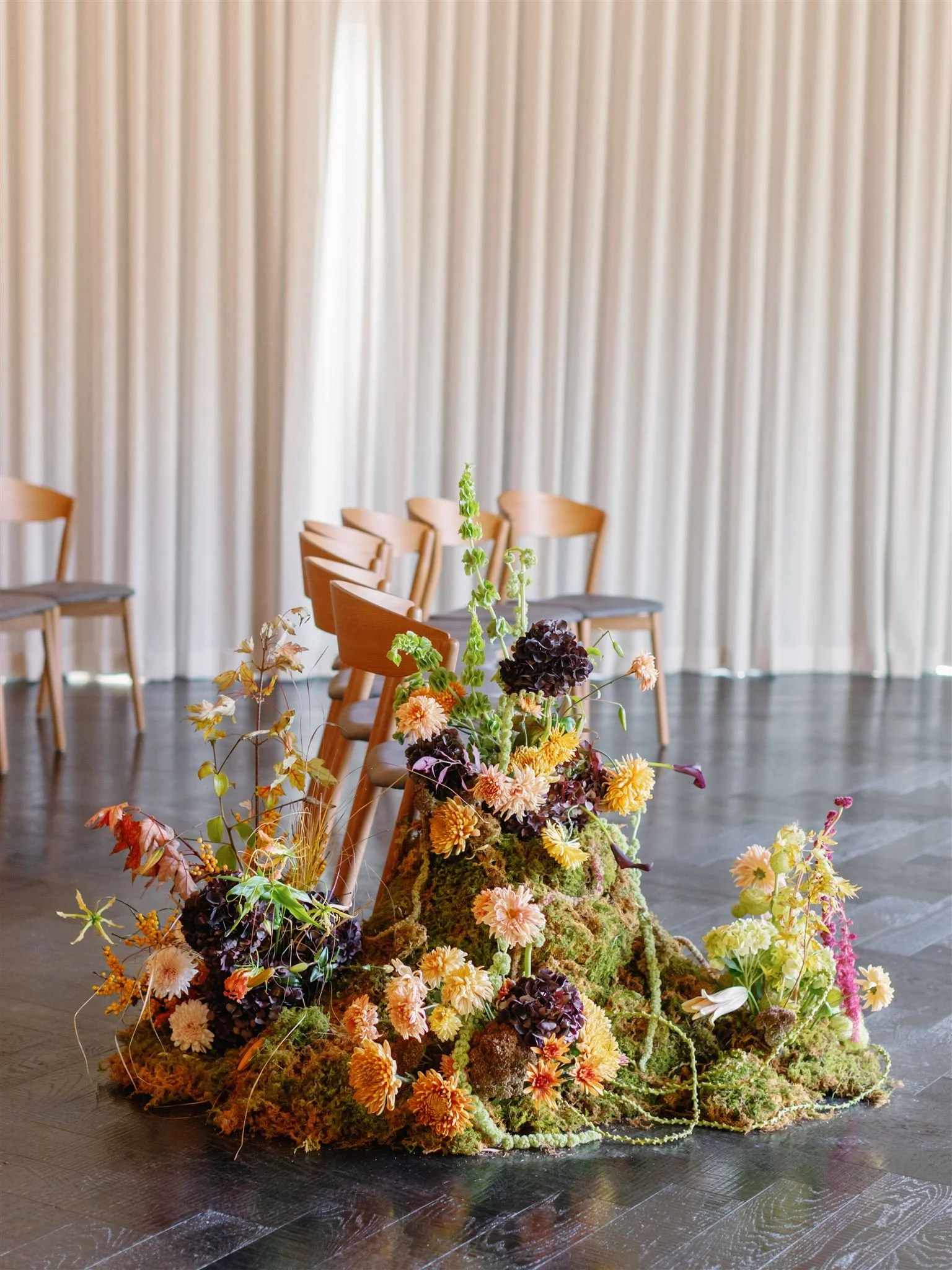 Indoor room with wooden chairs arranged in a semi-circle behind a floral arrangement on the floor, consisting of various flowers, moss, and greenery, positioned in front of a light-colored curtain.