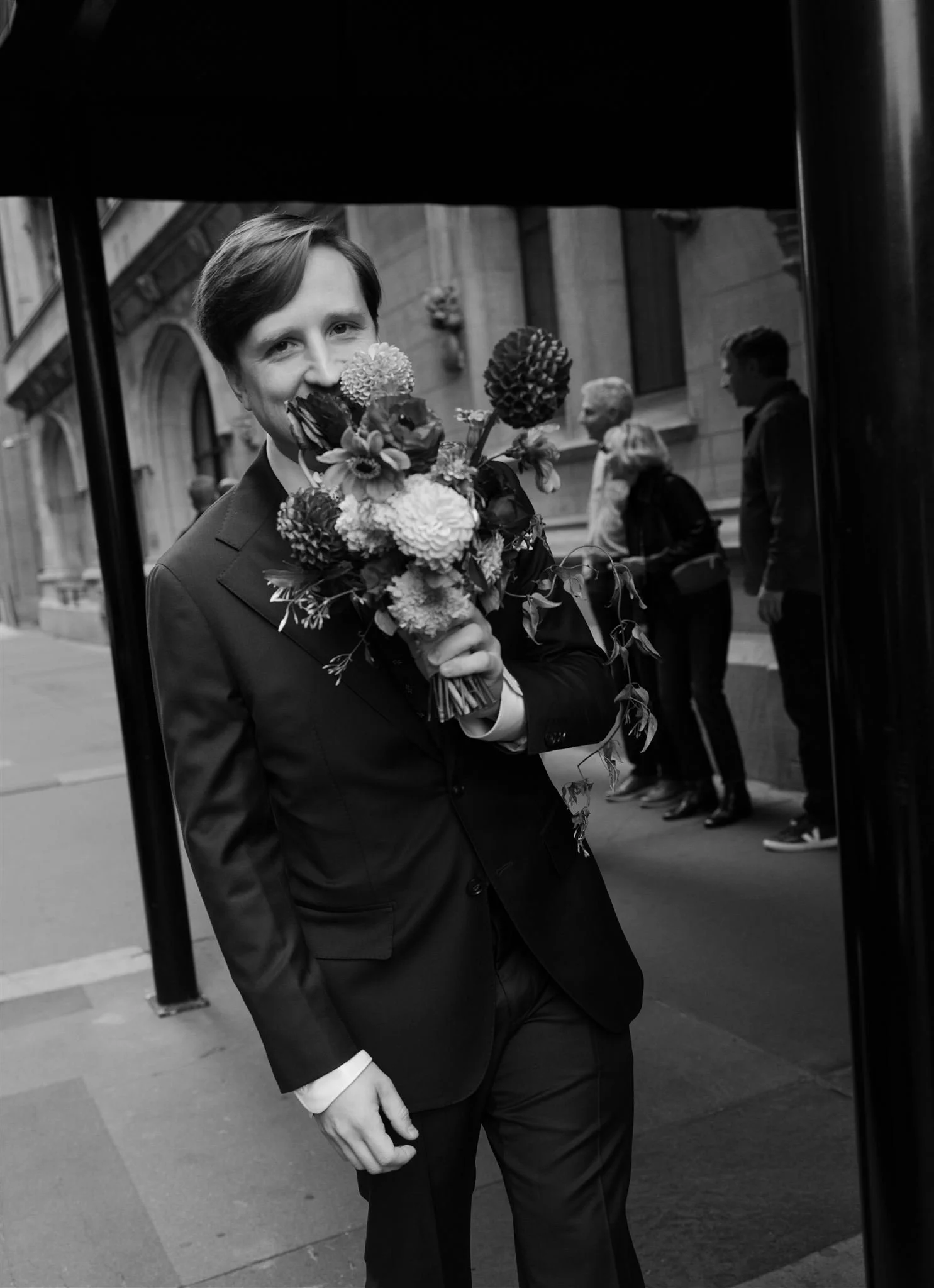 A person in a suit holding a bouquet of flowers, standing on a city sidewalk with a building and other pedestrians in the background.