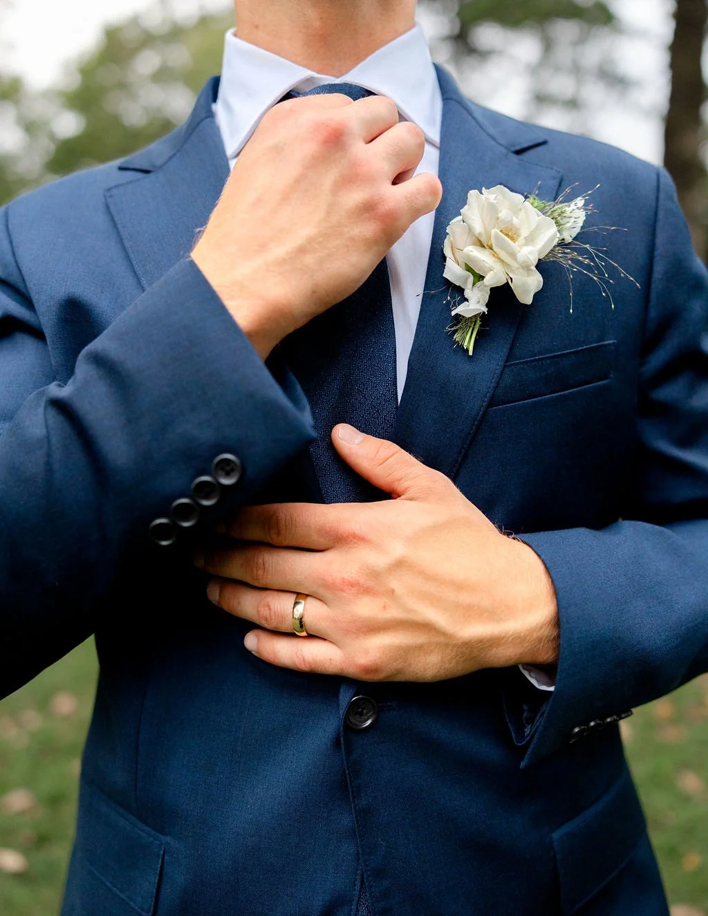 A man adjusting his blue suit tie with his left hand, wearing a wedding ring, with a white boutonniere on his lapel, outdoors.