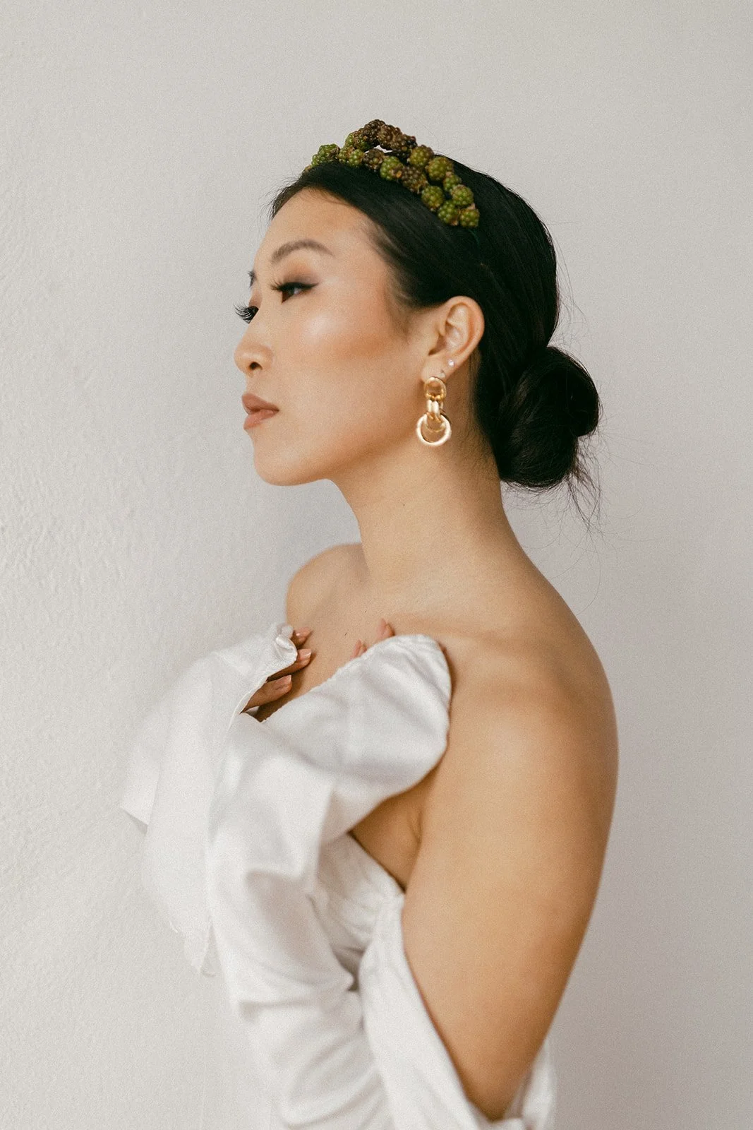 A woman with black hair styled in a low bun wearing white satin gloves, gold earrings, and a headpiece made of small green berries, posing against a plain white wall.