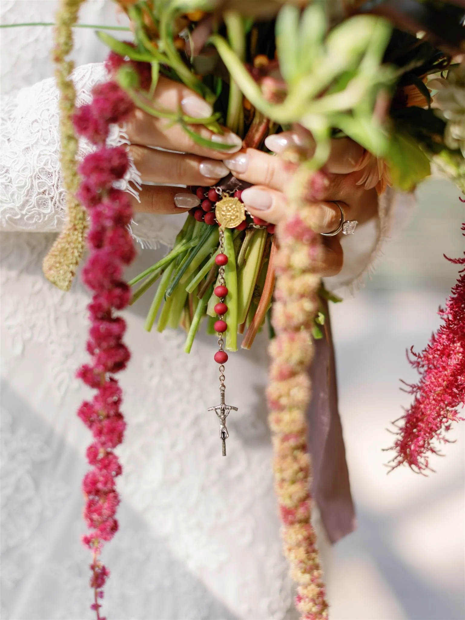 Close-up of a person holding a bouquet of flowers with a gold religious medal and a rosary with a crucifix. The person is wearing a white lace garment, and their hands display rings and a diamond ring.