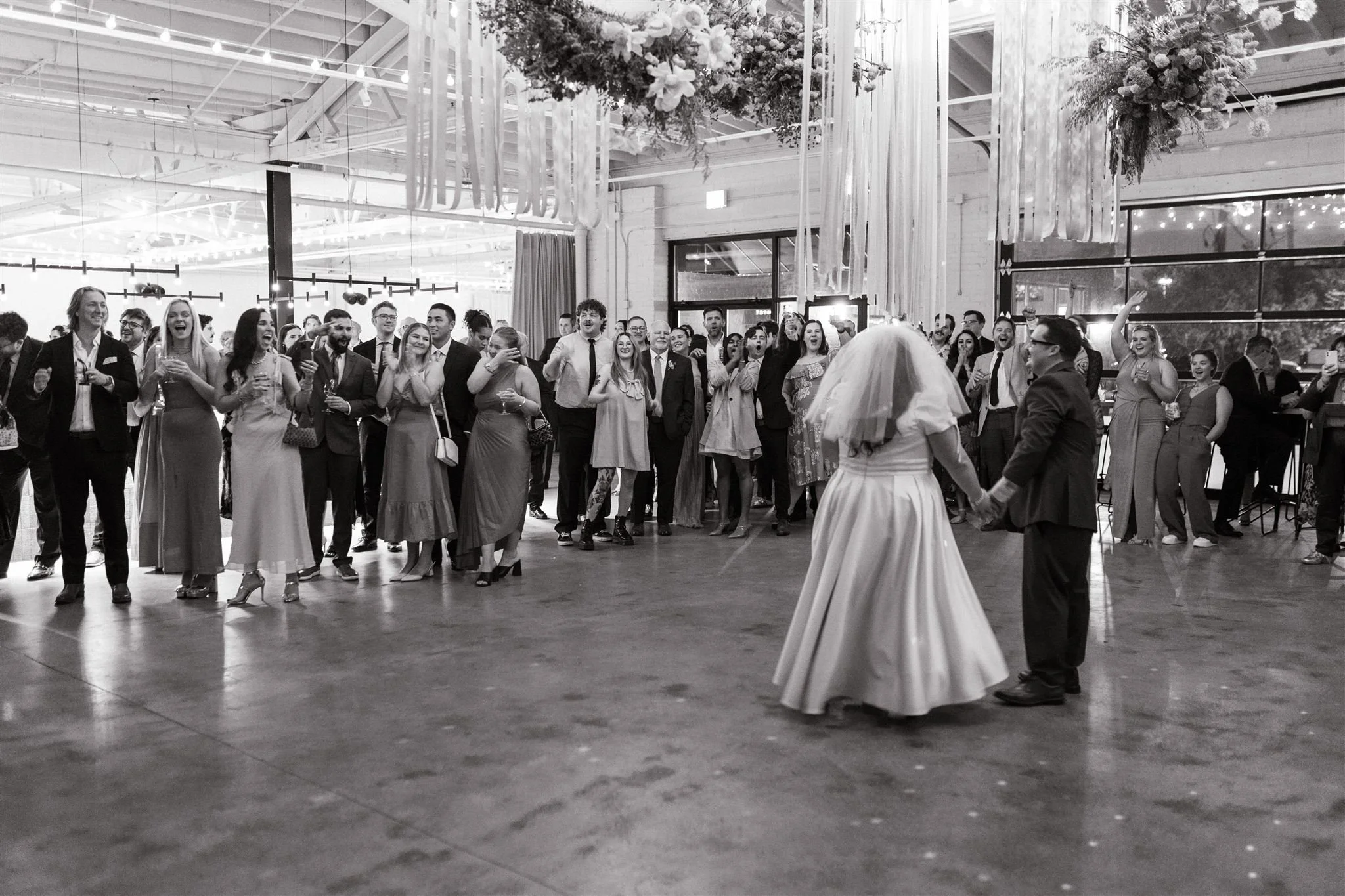 A black-and-white photo of a wedding reception. A bride and groom are dancing in front of a large crowd of guests in a decorated venue with floral arrangements and hanging ribbons.