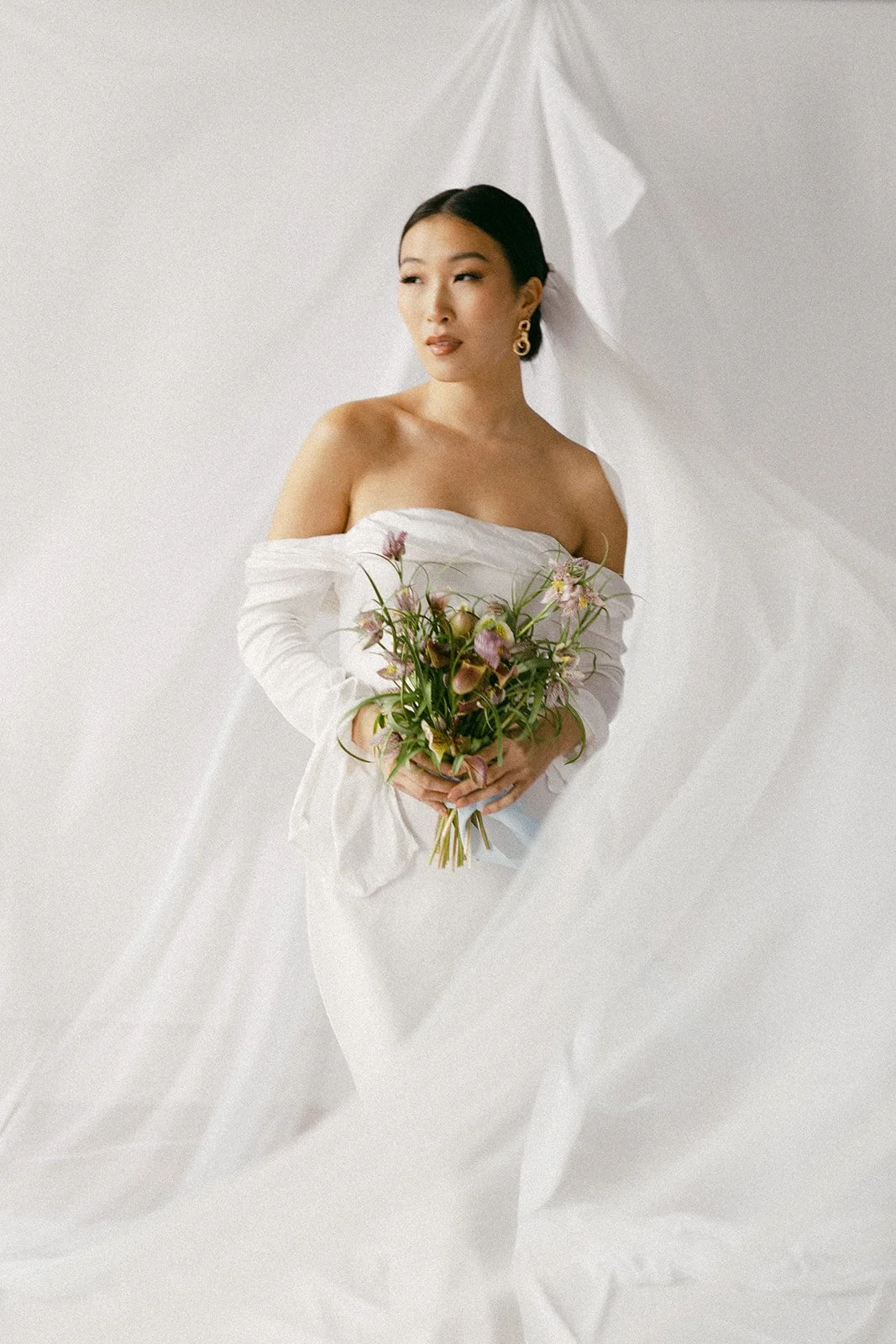 A woman in a white off-the-shoulder dress holding a bouquet of flowers, standing against a white background with draped fabric.