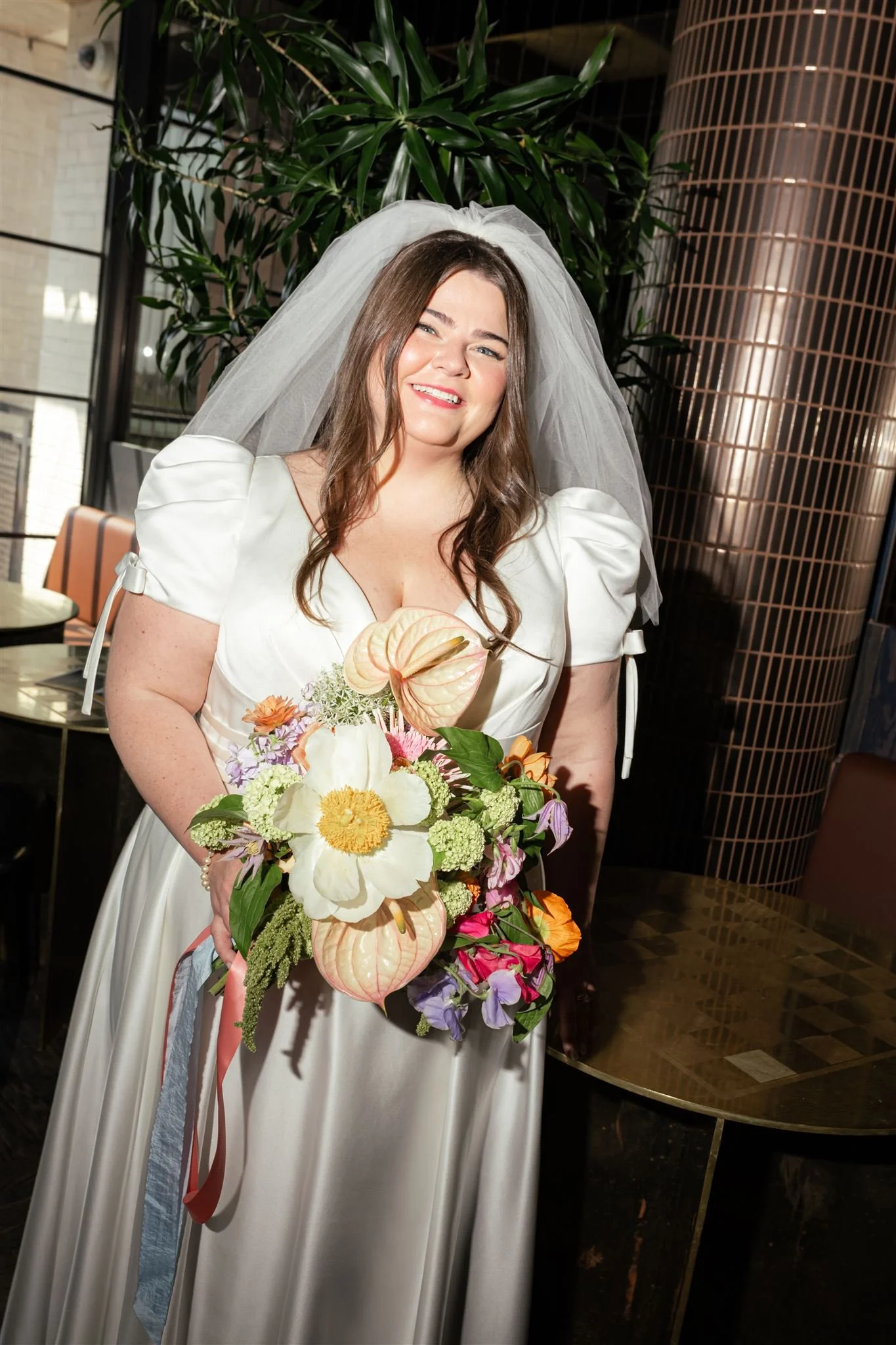 A woman in a white wedding dress and veil holding a bouquet of colorful flowers, smiling indoors with large green plants and modern decor in the background.