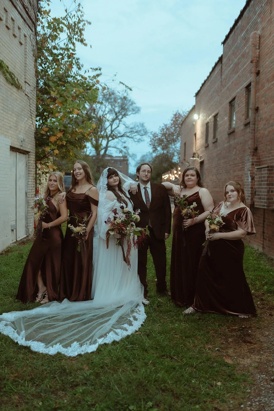 A wedding group portrait with a bride, groom, and four bridesmaids standing outdoors between brick buildings during early evening.