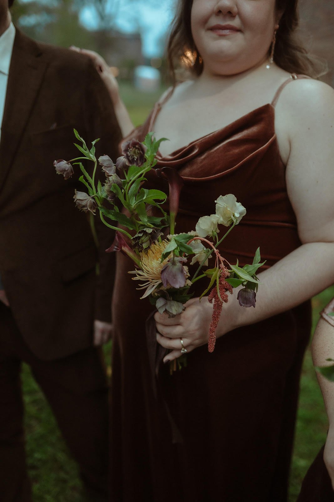 A woman in a brown velvet dress holding a bouquet of mixed flowers outdoors, with a man in a brown suit partially visible nearby.