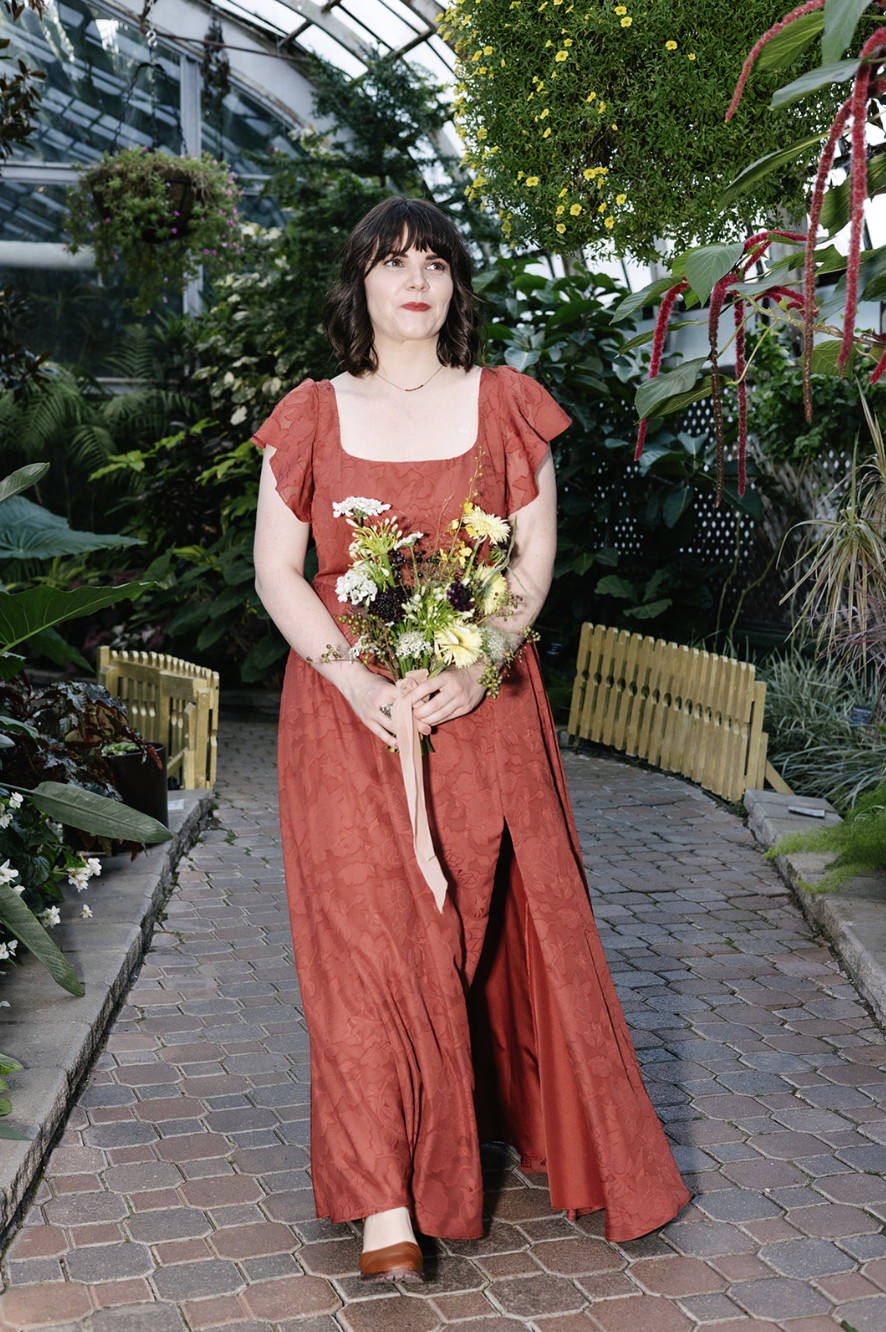 Woman in a red dress holding a bouquet of flowers, standing on a brick path inside a greenhouse surrounded by lush green plants and hanging flowers.