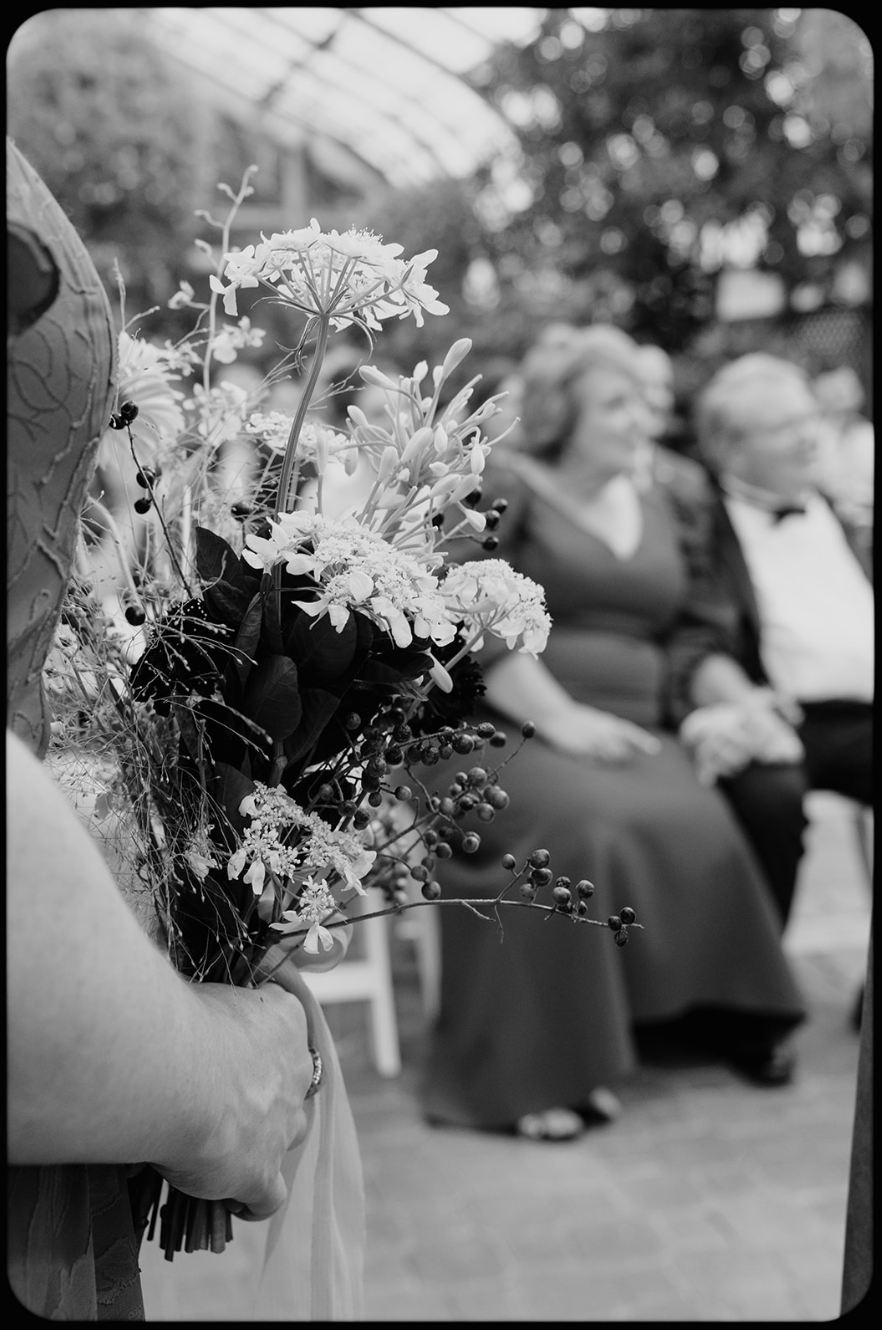 A woman holding a bouquet of flowers, with two women seated in the background at an outdoor event.