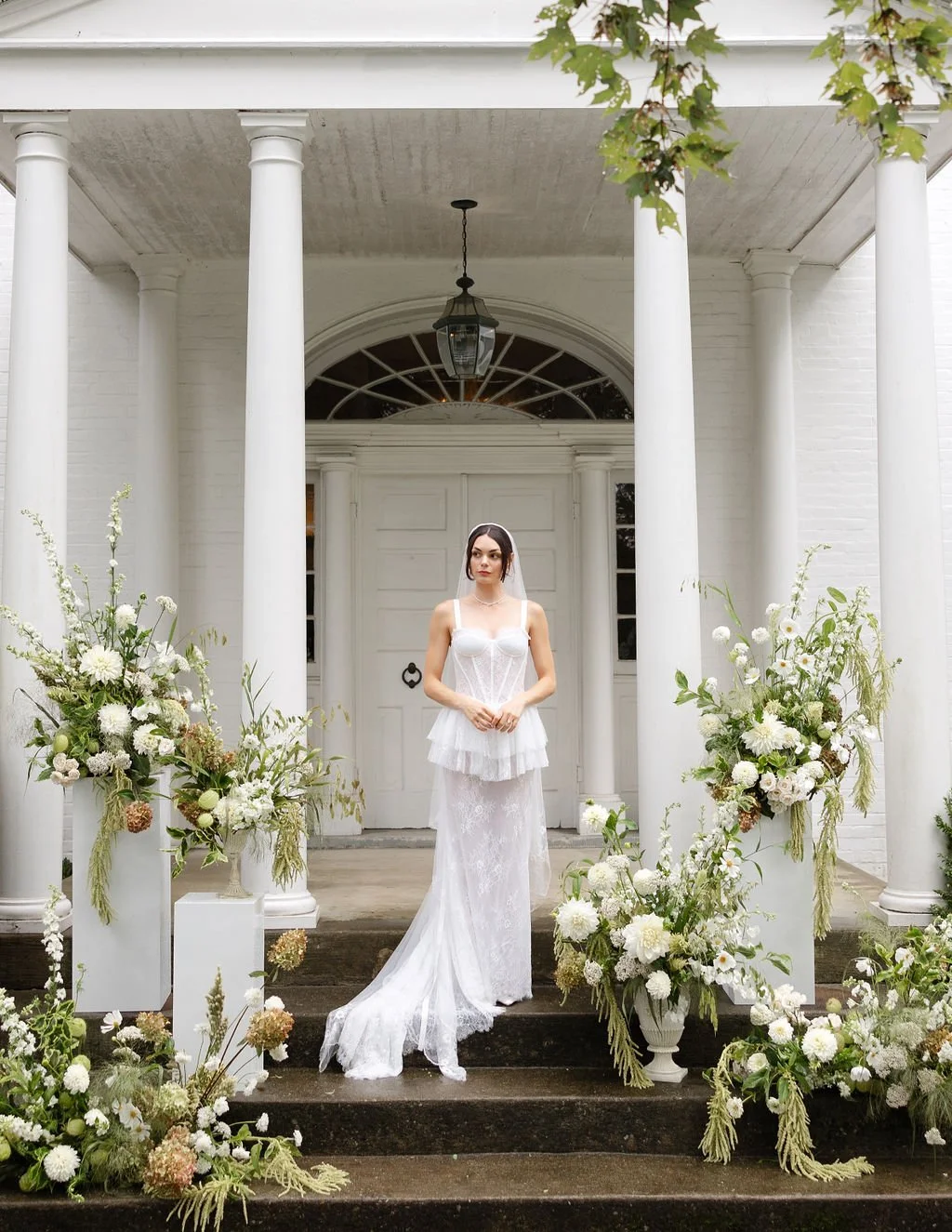 A bride in a white wedding dress standing on the steps of a white house decorated with large floral arrangements, white columns, and classical architectural elements.