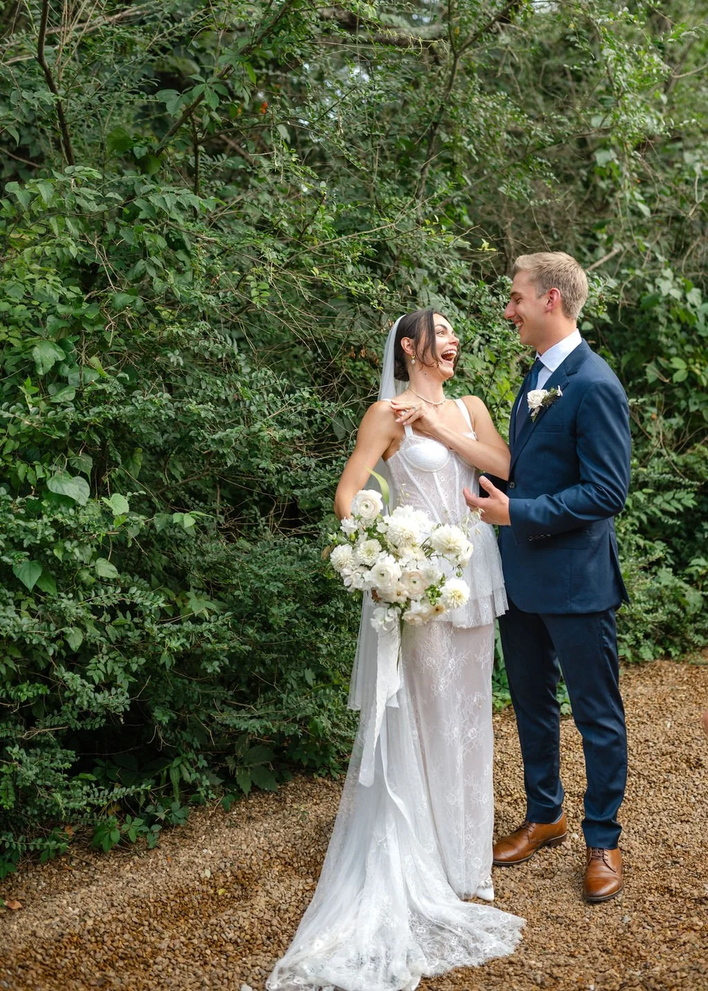 A bride and groom smiling and laughing together outdoors during their wedding, with lush greenery in the background. The bride is holding a bouquet of white flowers, and the groom is dressed in a navy suit with a tie and boutonniere.