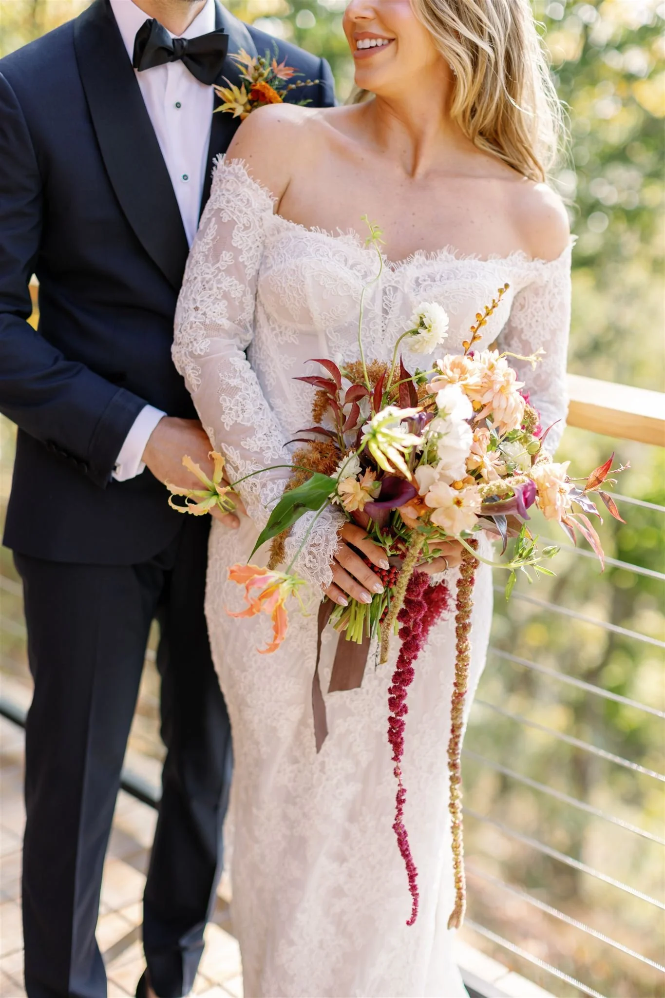 A bride in a white lace off-shoulder wedding gown holding a colorful bouquet of flowers, standing next to a groom in a dark suit with a bow tie, on a wooden deck with a forest background.