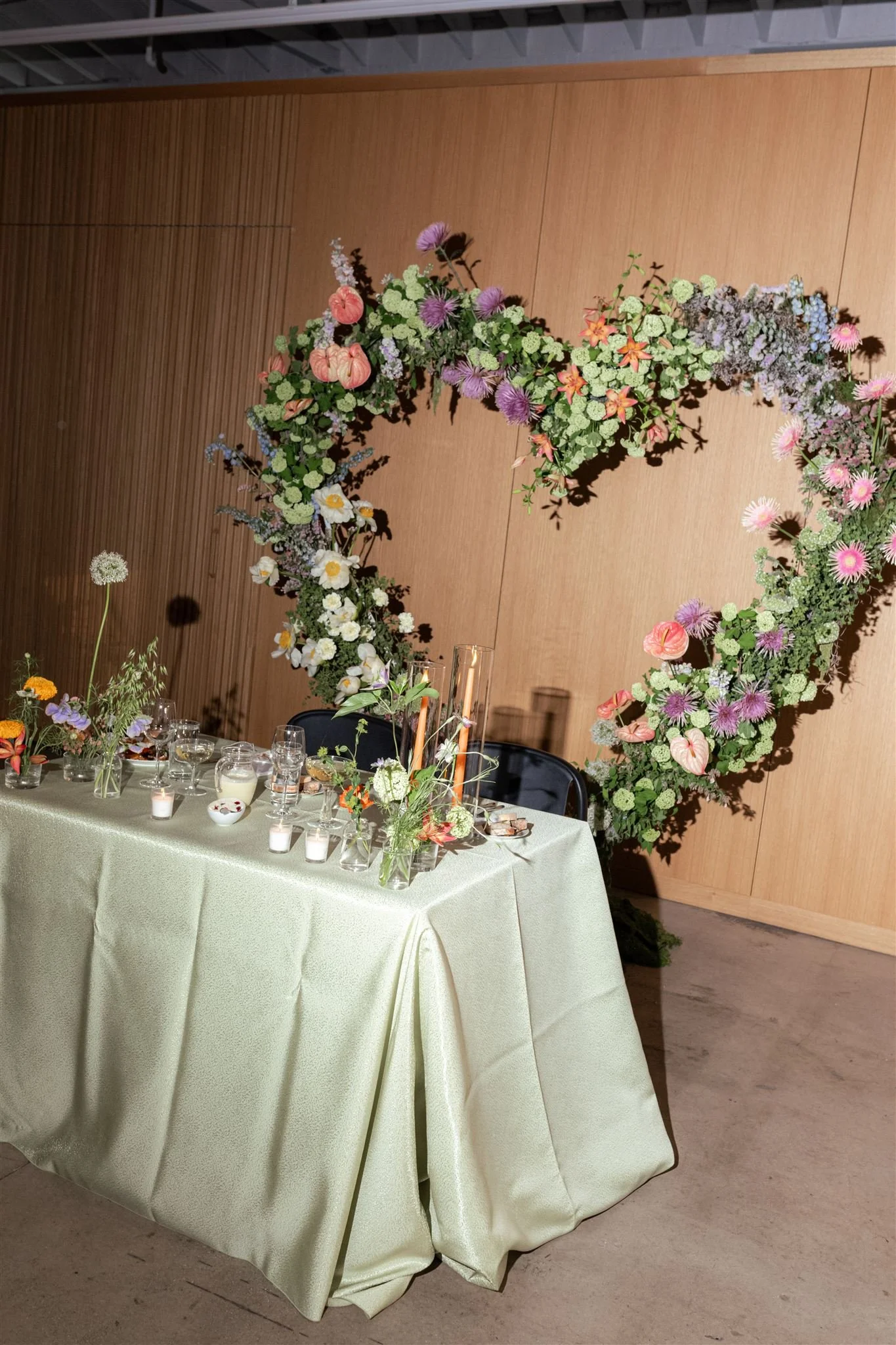 A decorated table with candles, vases, and flowers in front of a large heart-shaped floral wreath on a wood-paneled wall, likely for a wedding or celebration.