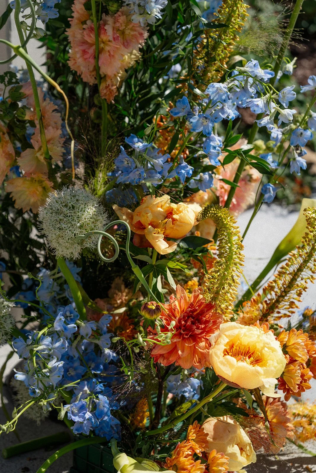 A vibrant floral arrangement with blue delphinium, yellow and peach carnations, orange dahlias, and various greenery.