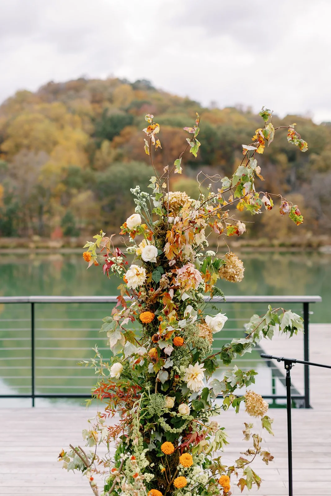 Flower arrangement with various autumn-colored flowers and leaves, outdoors near a lake with a tree-covered hill in the background.