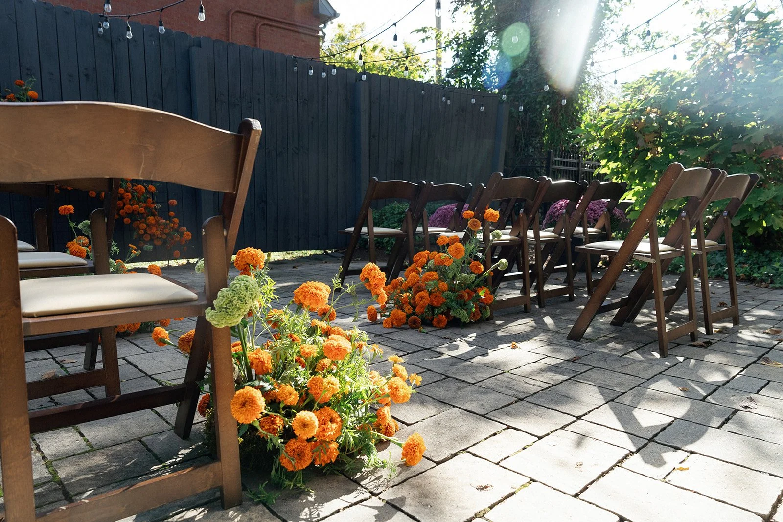 Outdoor patio with wooden chairs, orange and green flowers, string lights, and a blue fence in sunlight.