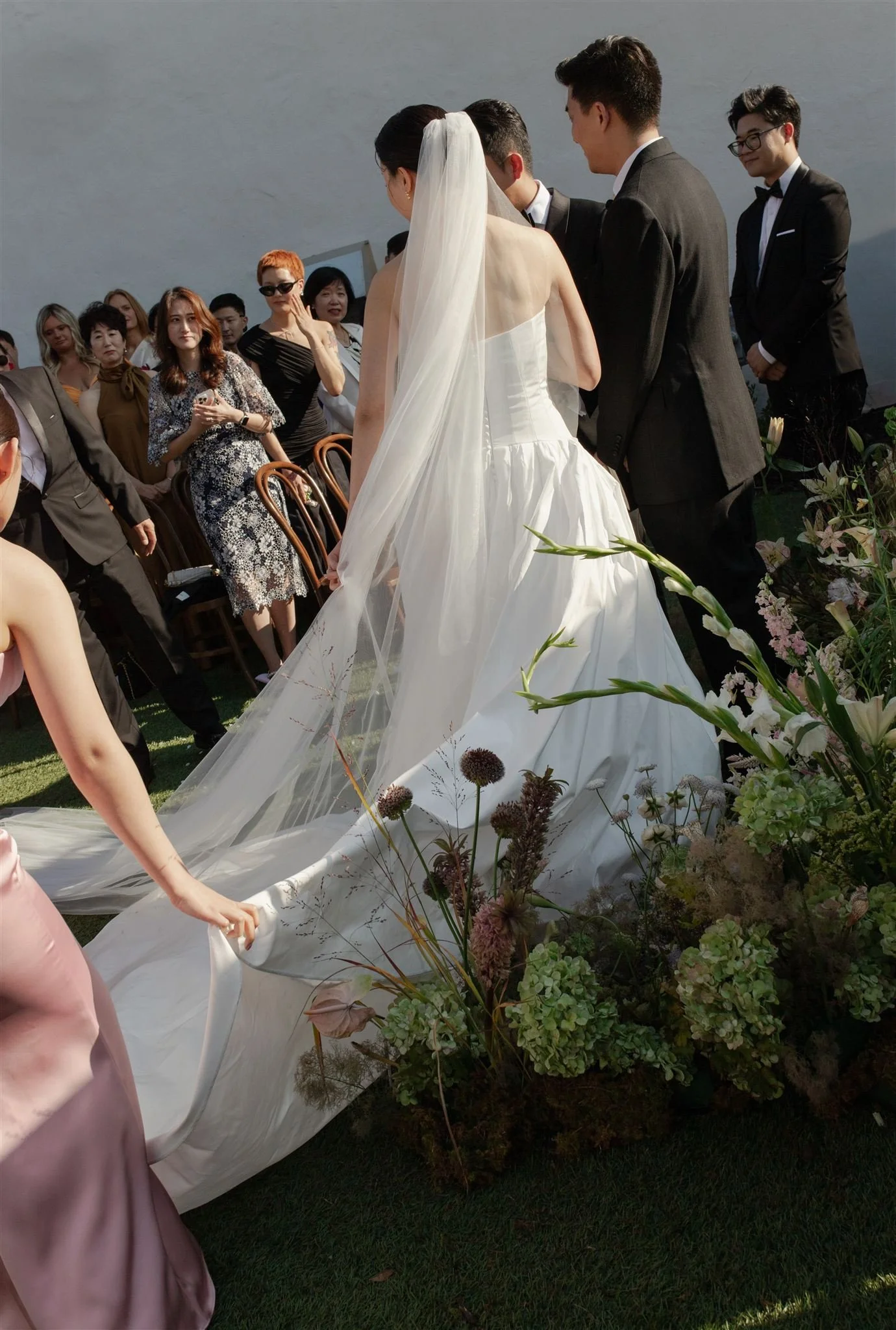 A bride in a white wedding gown with a long train and veil, standing outdoors with a groom and guests attending a wedding ceremony.