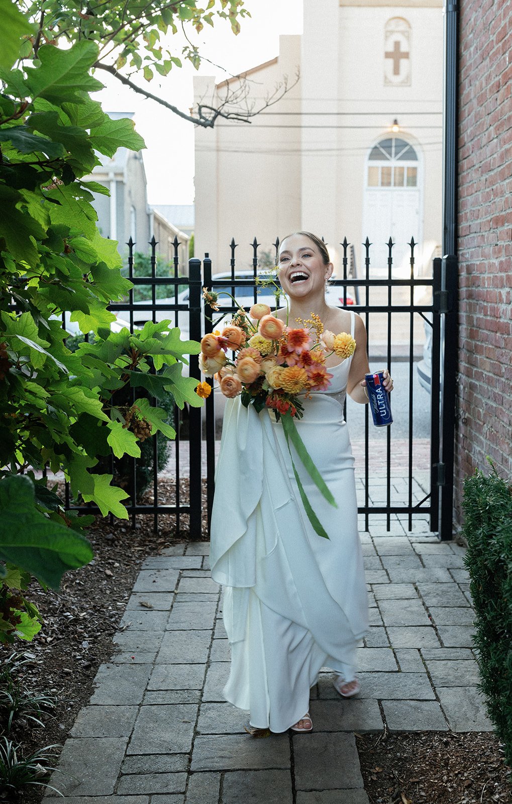 A smiling woman in a white dress holding a large bouquet of orange and yellow flowers, with a can of Ultra beer in her right hand, walking on a paved pathway near a brick wall and green bushes.