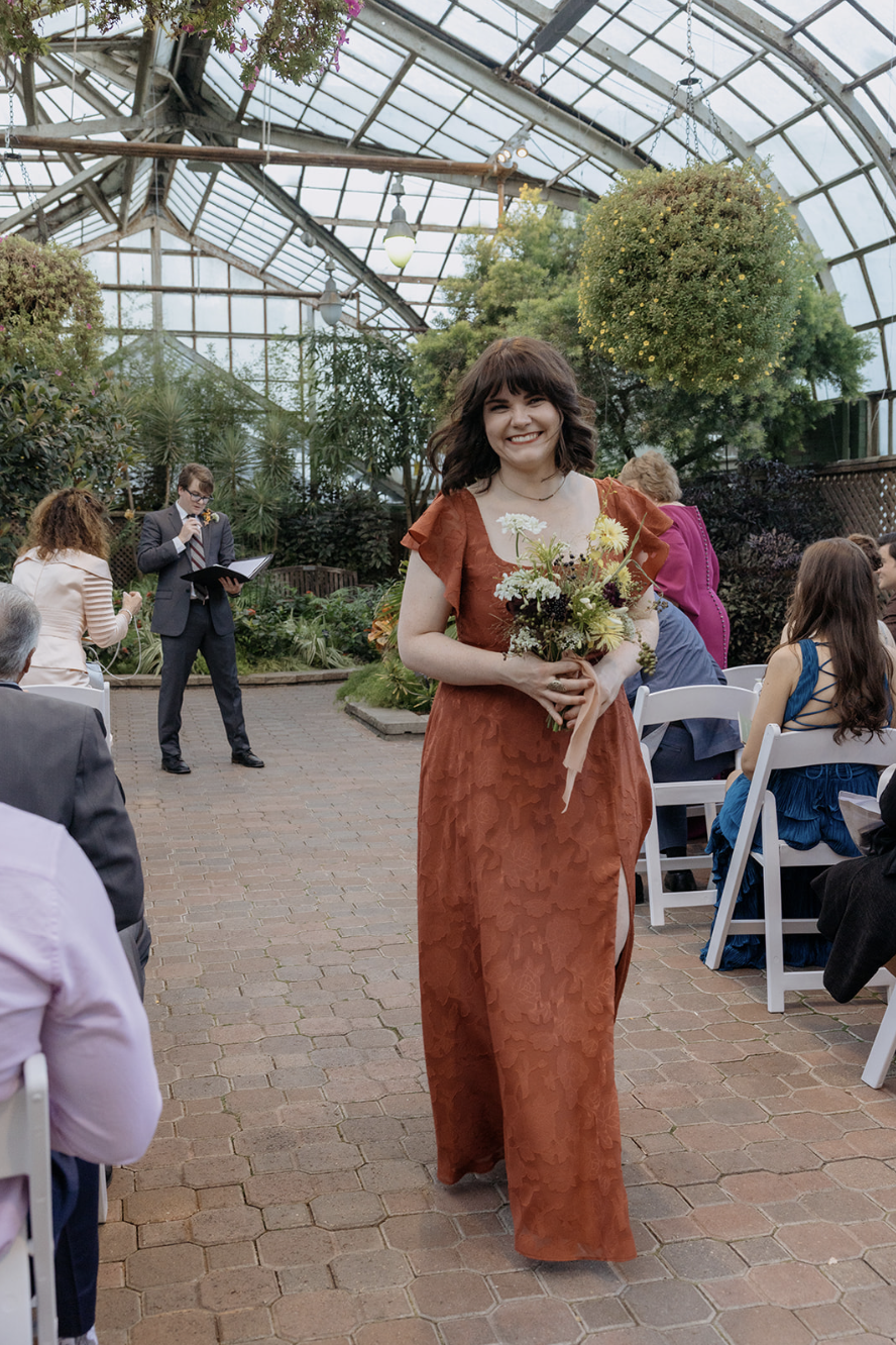 A smiling woman in a rust-colored dress holding a bouquet of flowers at an indoor garden event surrounded by seated guests and a man in a suit reading from a book in the background.