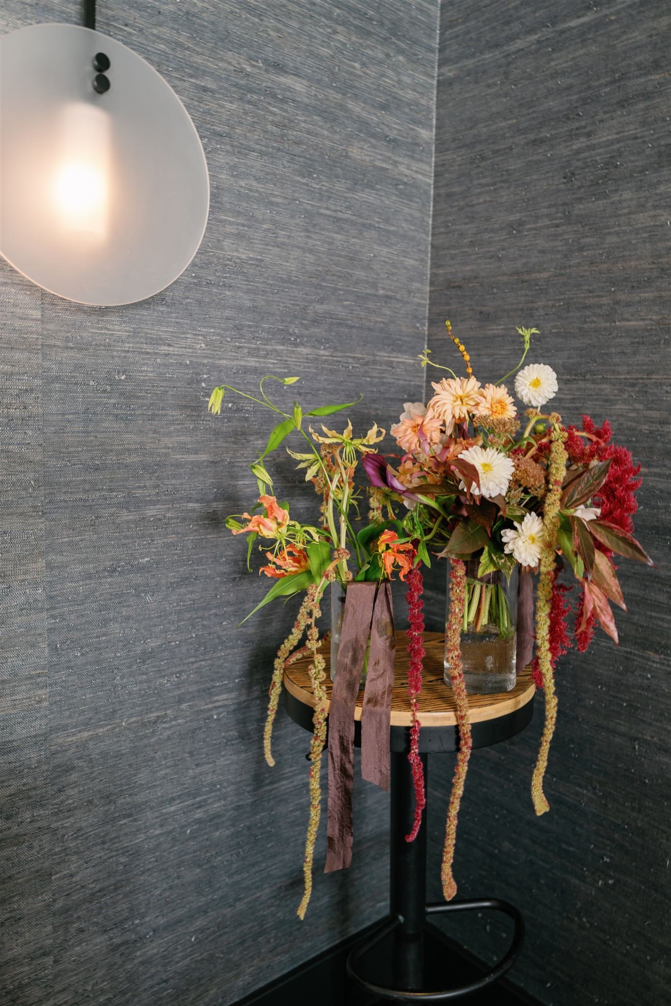 A floral arrangement with pink, white, red, and purple flowers in a glass vase on a small round wooden table against a textured dark gray wall, with a modern wall sconce above.