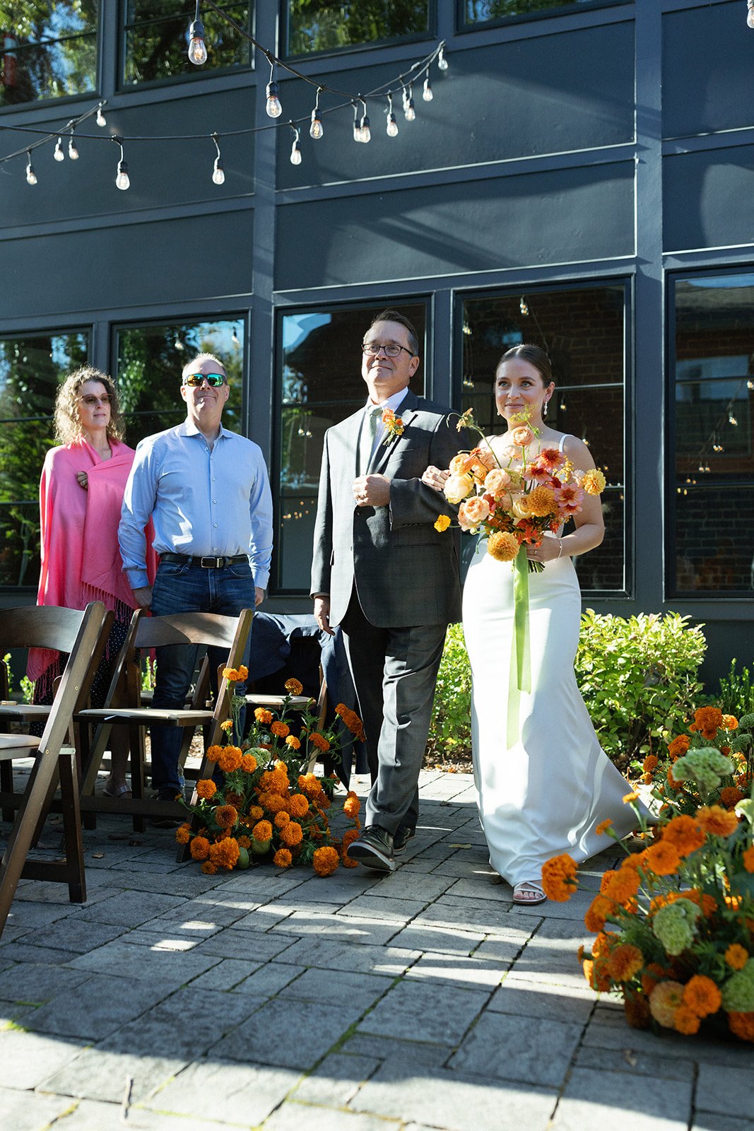 A bride in a white wedding dress holding a bouquet of orange and yellow flowers, walking down the aisle with a man in a suit at an outdoor wedding ceremony. Two other guests stand in the background, under string lights hanging above, with orange flow