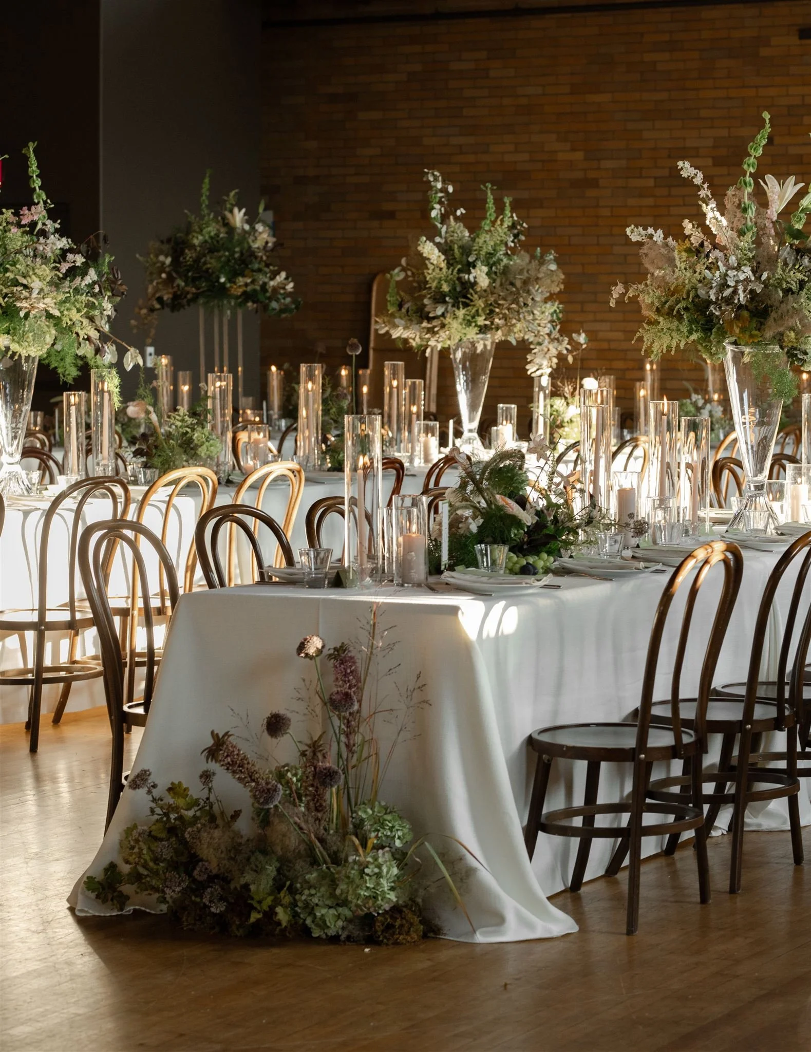 Elegant banquet table with white tablecloths, tall candle holders, and floral centerpieces with greenery and white flowers, set against a brick wall.